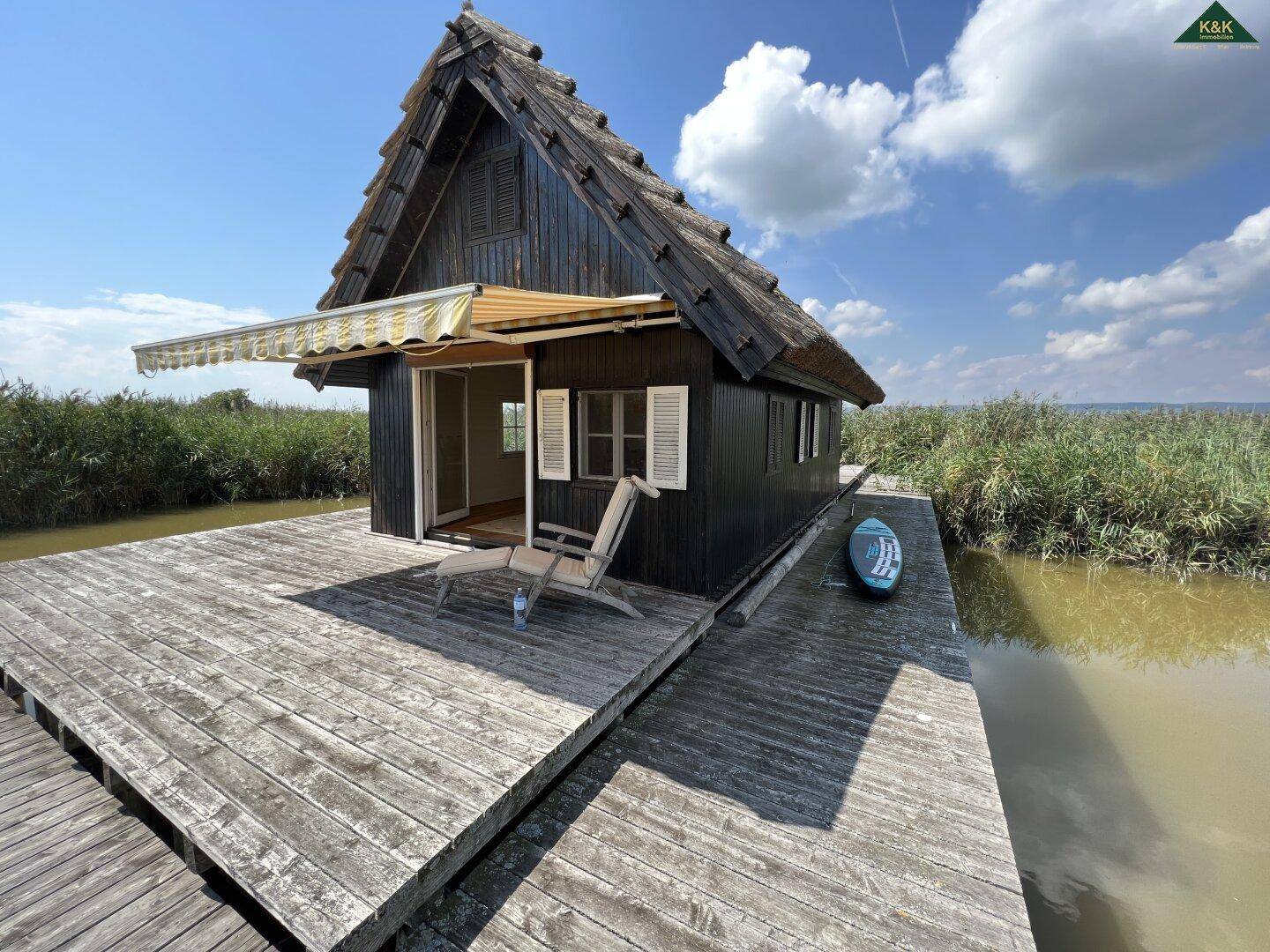 Charmantes Holzhaus auf einem weitläufigen Holzsteg, umgeben von Natur und Wasser, unter blauem Himmel.