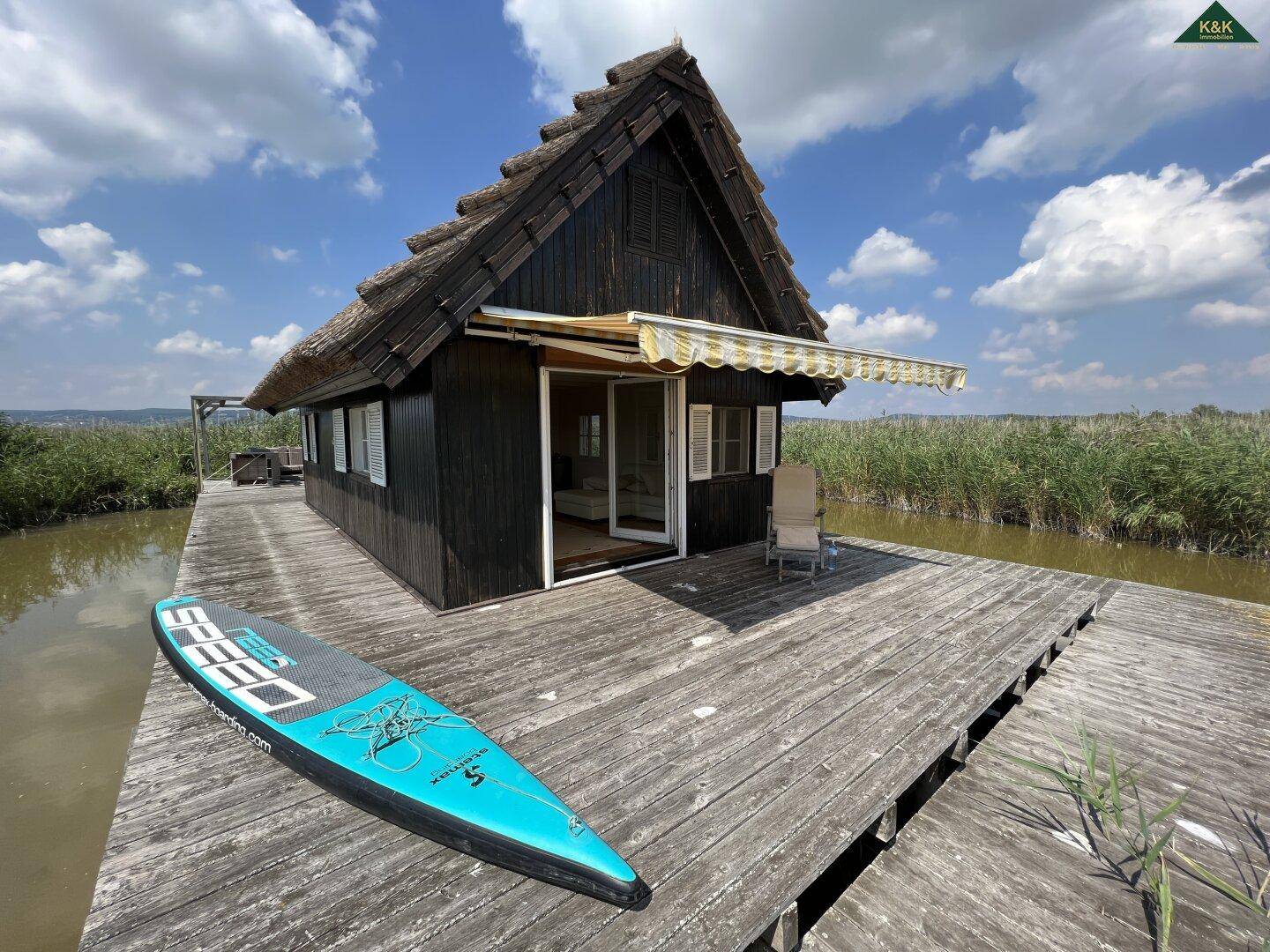 Charmantes Holzhaus auf einem Holzsteg über dem Wasser, umgeben von Schilf, mit einem Paddleboard.
