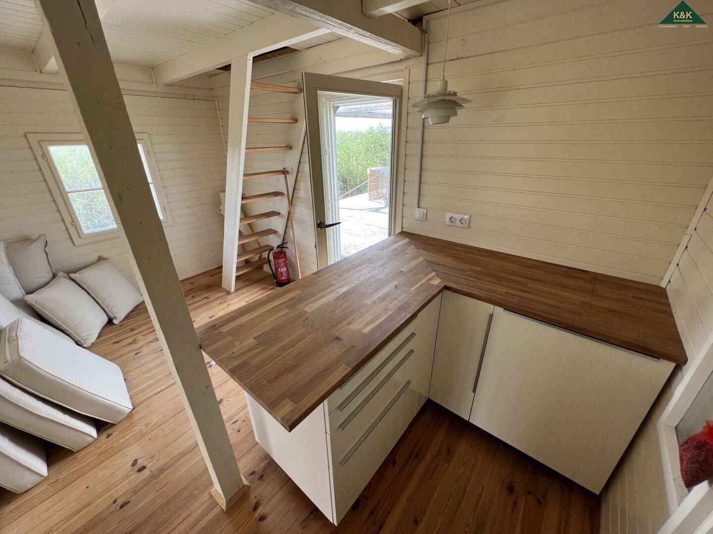 Kleine Küche mit weißen Unterschränken und Holz-Arbeitsplatten, daneben eine steile Holztreppe zum Obergeschoss.