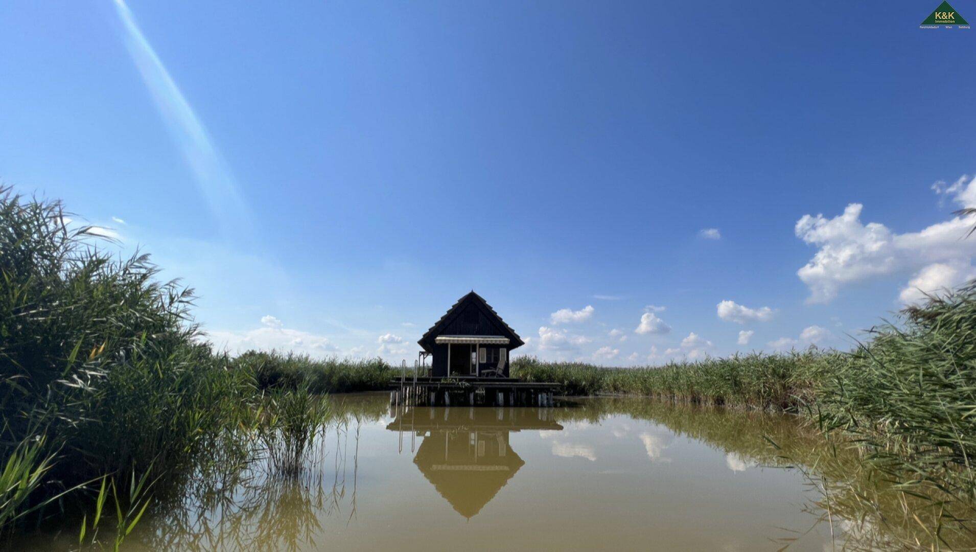 Malerisches Holzhaus inmitten von Schilf und Wasser, spiegelt sich im ruhigen See unter blauem Himmel.