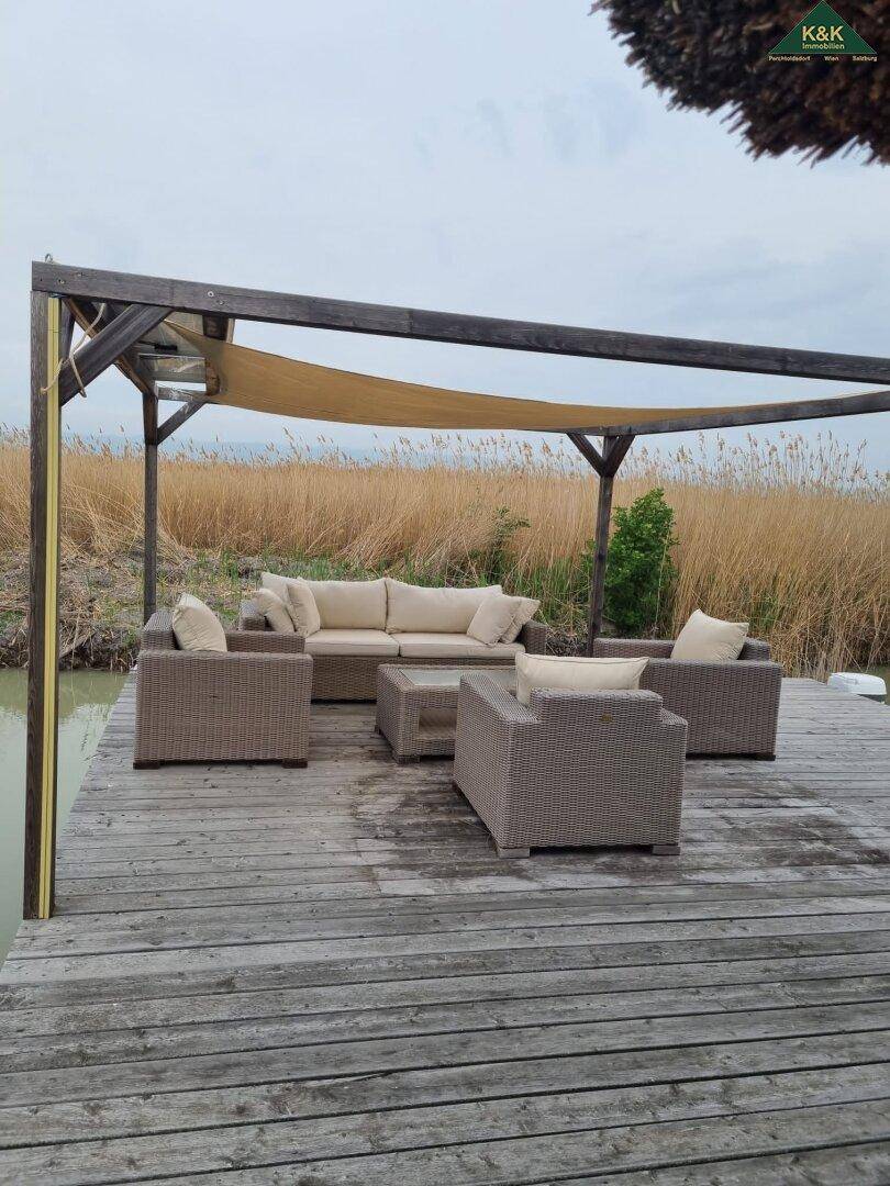 Möblierter Außenbereich auf einem Holzsteg mit Pergola und Blick auf das umgebende Schilf und Wasser.