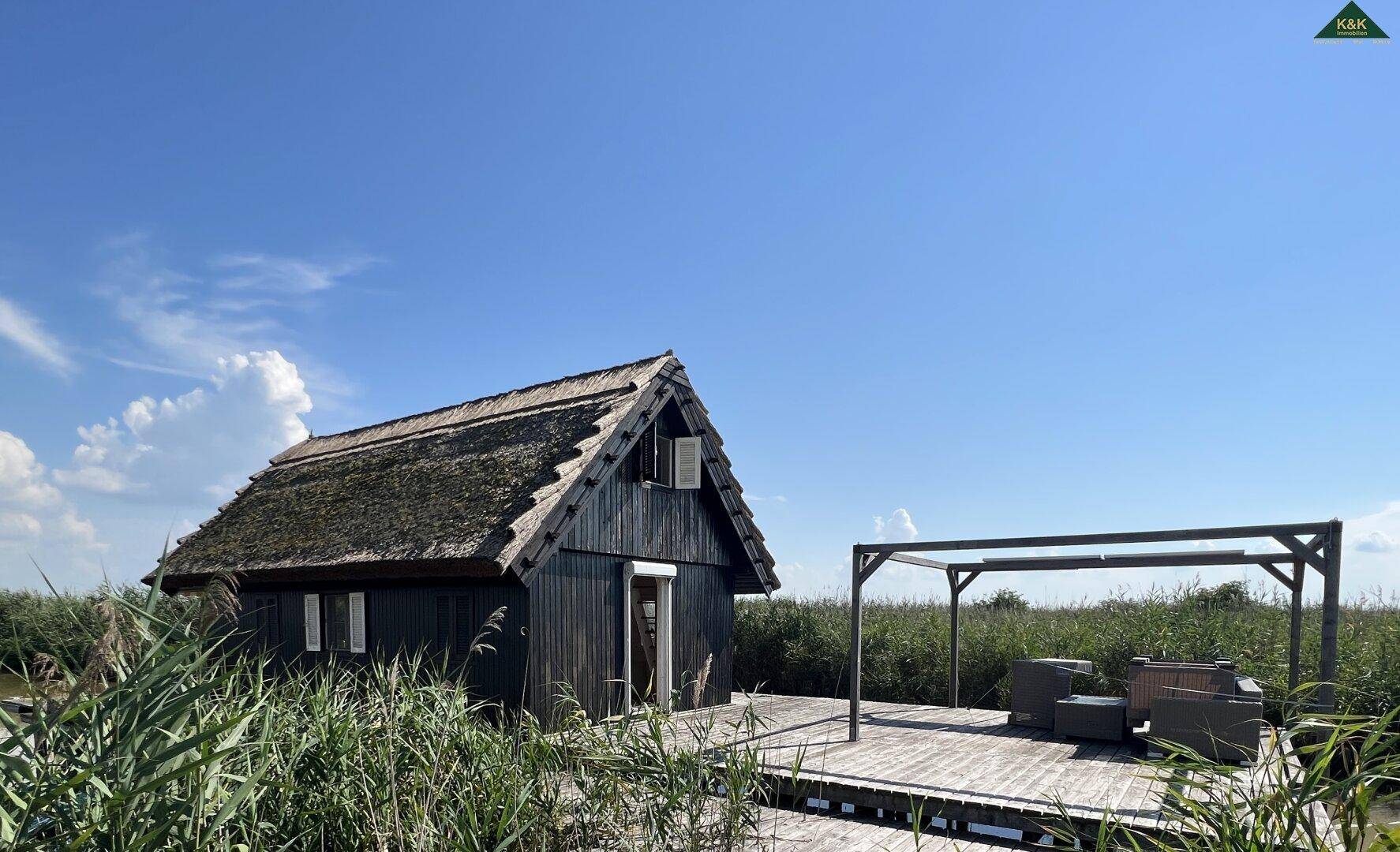 Kleines Holzhaus mit Reetdach und einer Holzterrasse, umgeben von Schilf unter blauem Himmel.