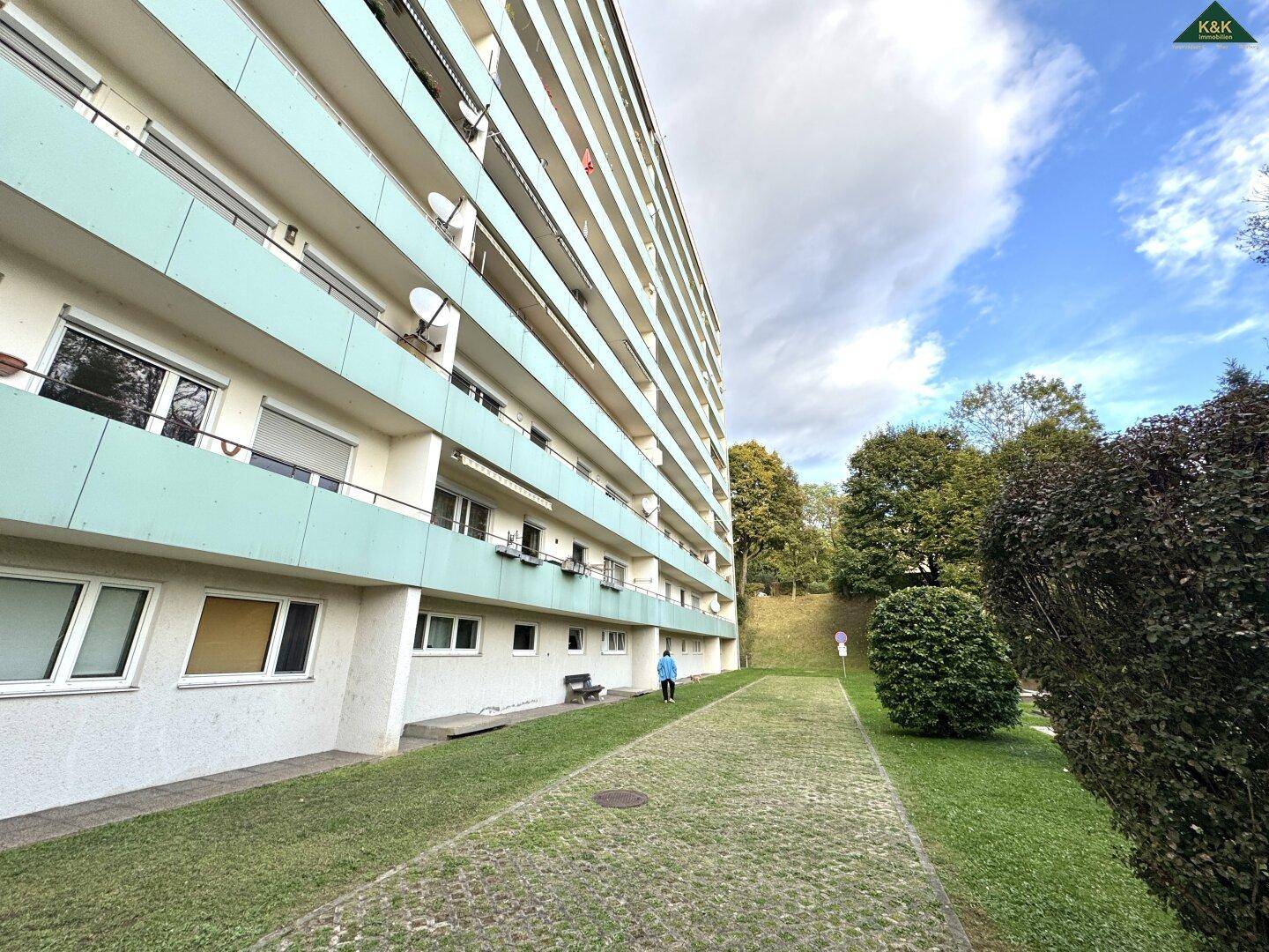 Seitenansicht eines Mehrfamilienhauses mit Balkonen und Grünflächen, unter bewölktem Himmel.