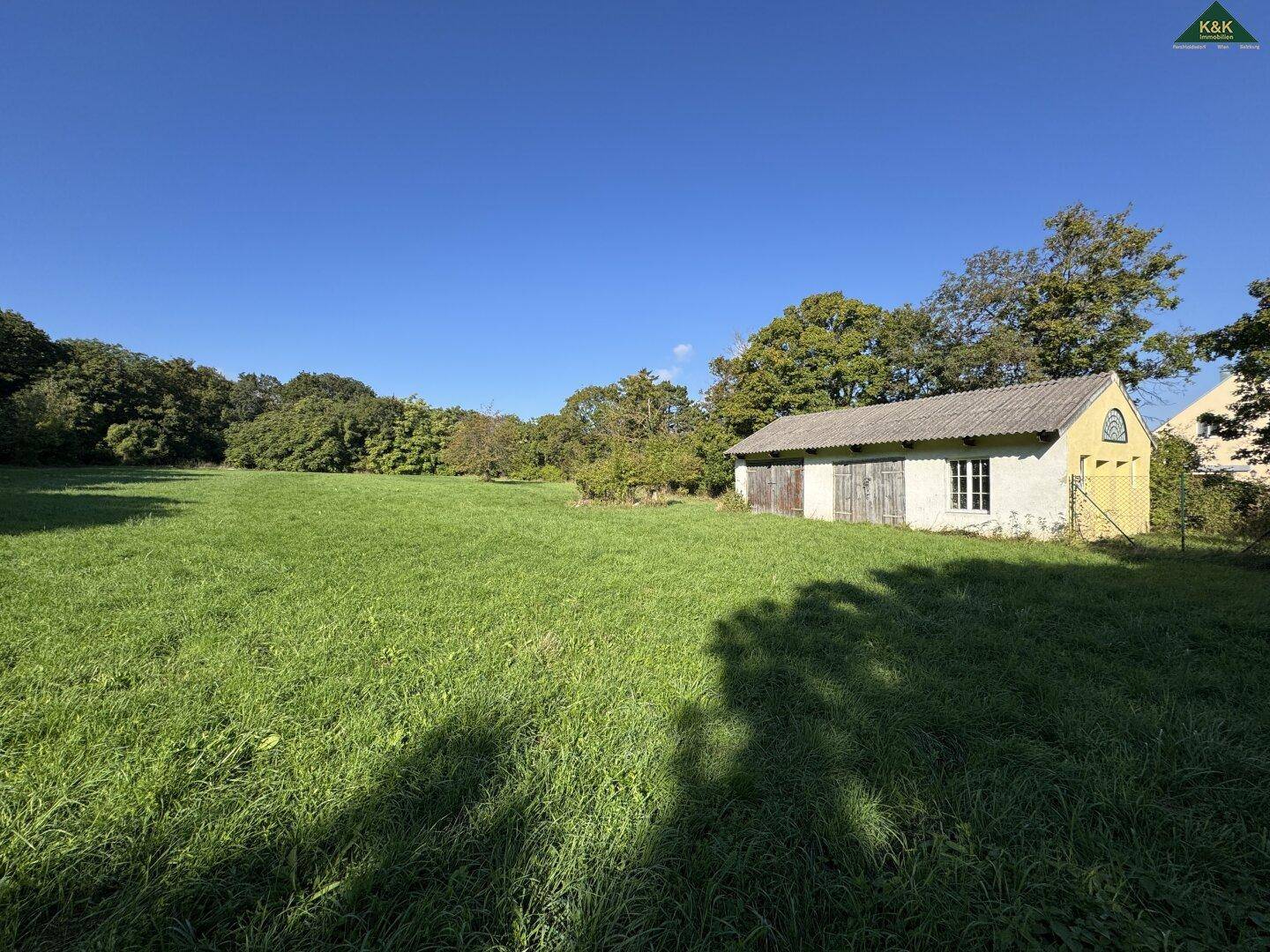 Weitläufige grüne Wiese mit einem kleinen Nebengebäude im Hintergrund unter blauem Himmel.