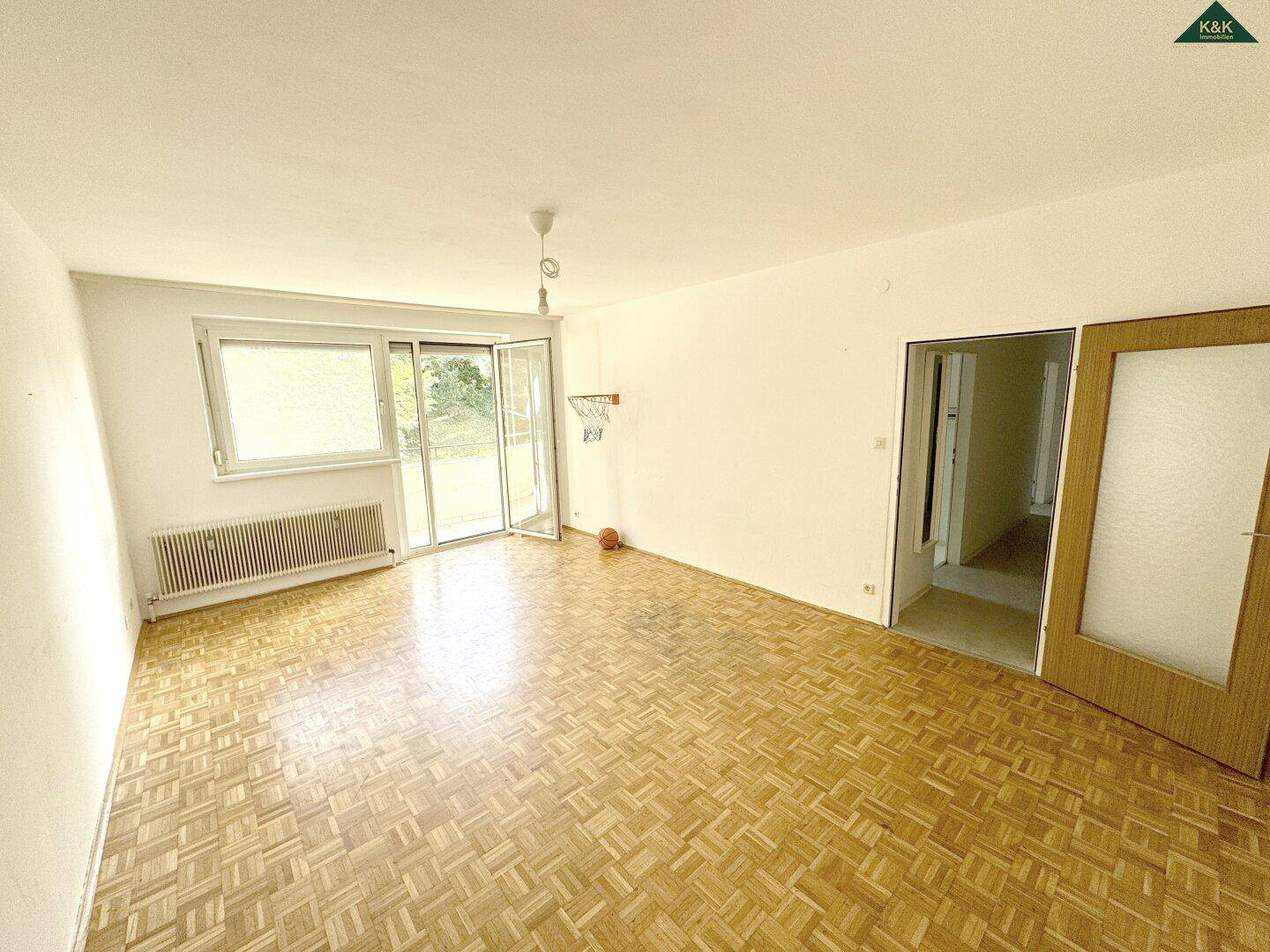 Geräumiges Zimmer mit Fischgrätparkett, großem Fenster und Zugang zu einem Außenbereich.