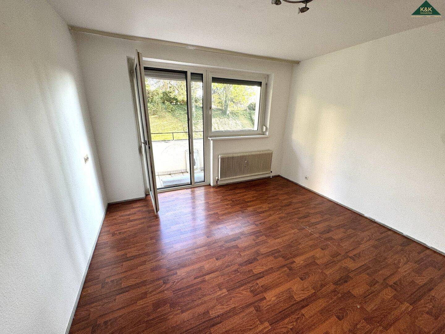 Lichtdurchflutetes Zimmer mit dunklem Laminatboden und direktem Zugang zum Balkon.