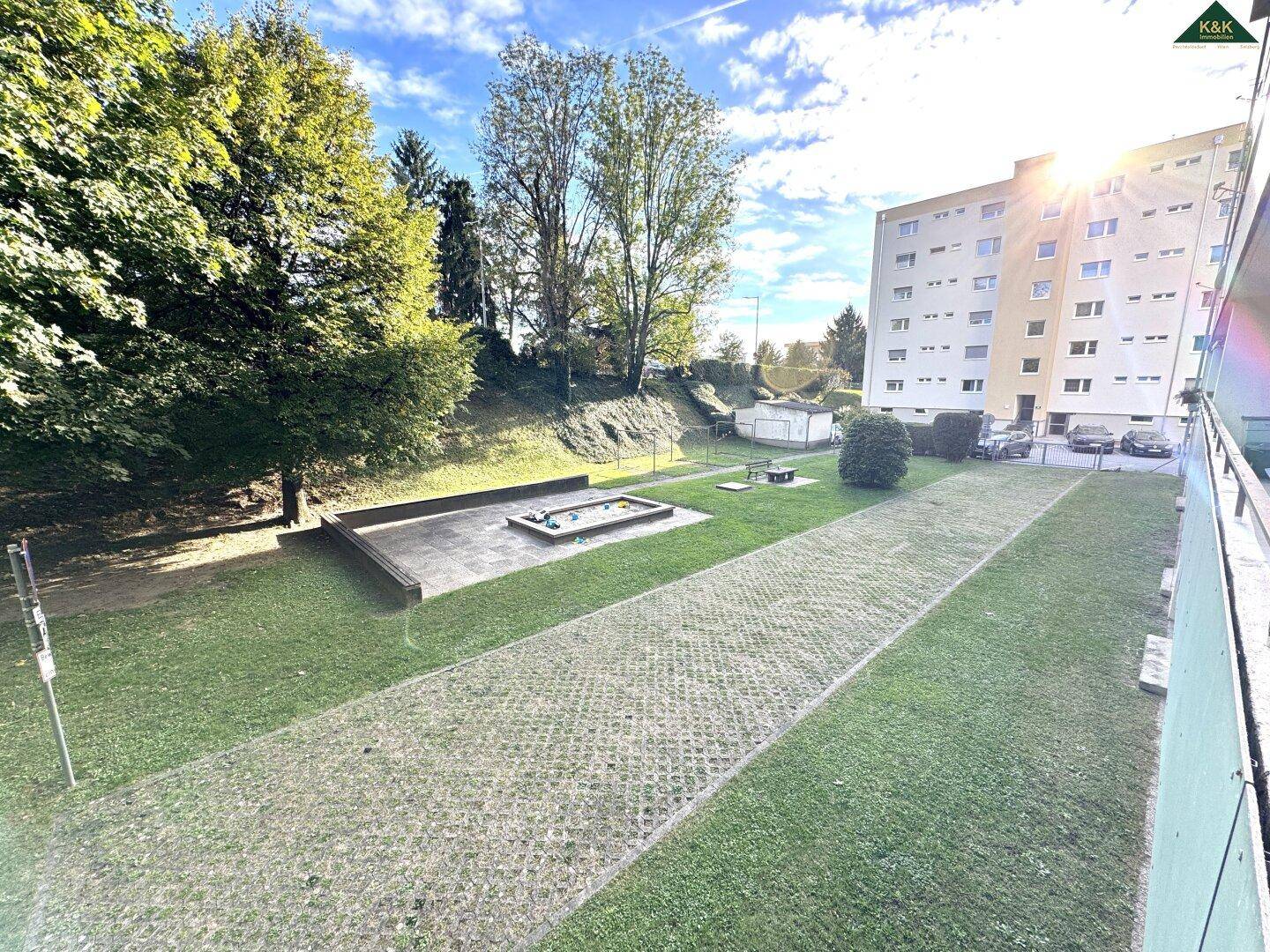 Gemeinschaftsgarten mit Rasenflächen, Bäumen und einem kleinen Spielbereich unter blauem Himmel.