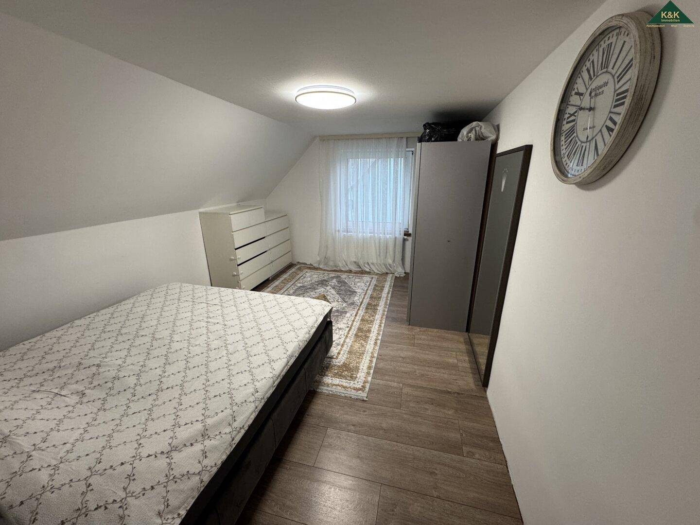 Helles Schlafzimmer im Dachgeschoss mit schrägen Wänden und einem großen Wanduhr.