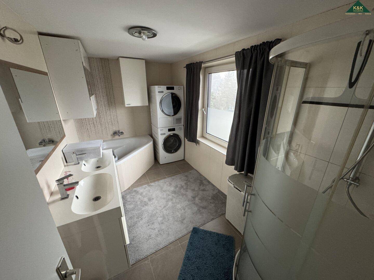 Funktionales Badezimmer mit Eckbadewanne, Dusche, Doppelwaschbecken und Fenster, inklusive Waschmaschine und Trockner.