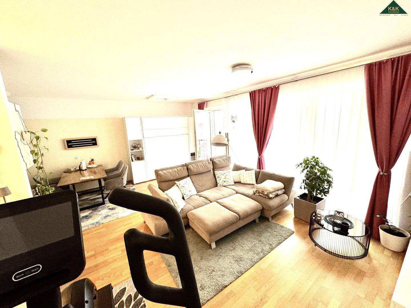 Helles Wohnzimmer mit bequemer Couch, Essbereich und großen Fenstern mit roten Vorhängen.
