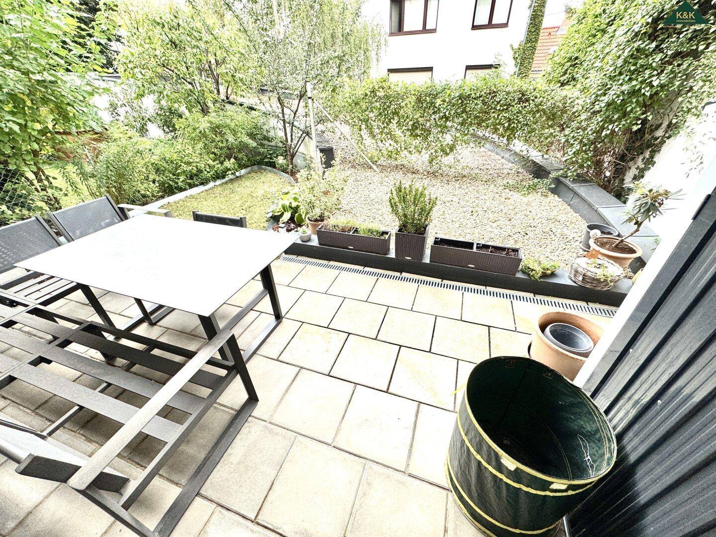 Heller Terrassenbereich mit Sitzgelegenheiten und Zugang zum Garten, umgeben von üppigem Grün.