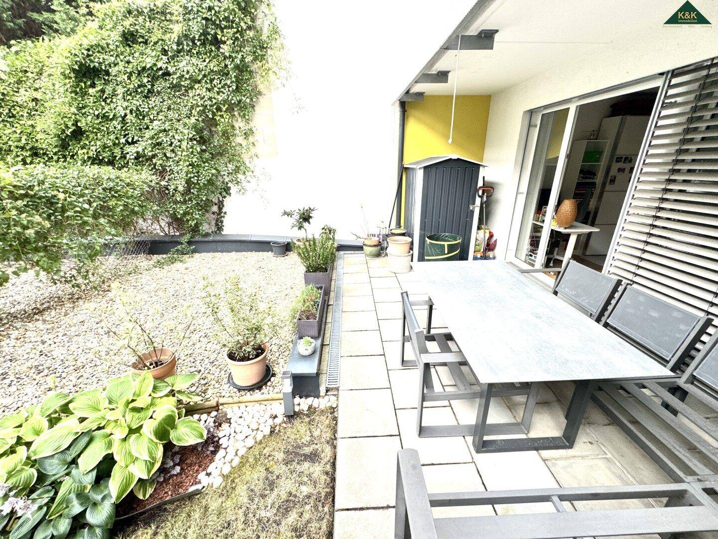 Heller Terrassenbereich mit Sitzgelegenheiten und Zugang zum Garten, umgeben von üppigem Grün.