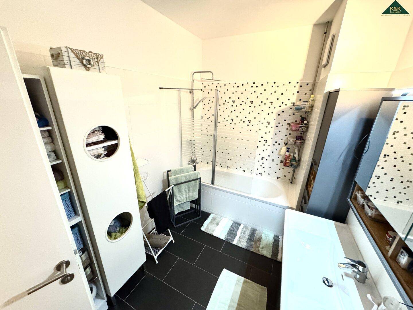 Badezimmer mit Badewanne und Duschwand, ausgestattet mit dunklen Fliesen und Stauraum.