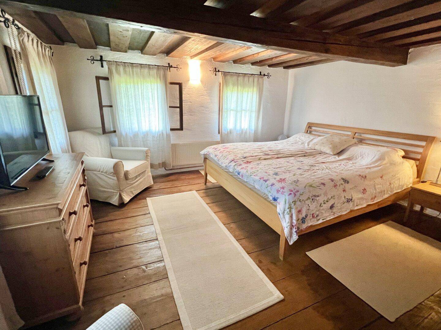Gemütliches Schlafzimmer mit Holzboden, sichtbaren Deckenbalken und zwei Fenstern für natürliches Licht.