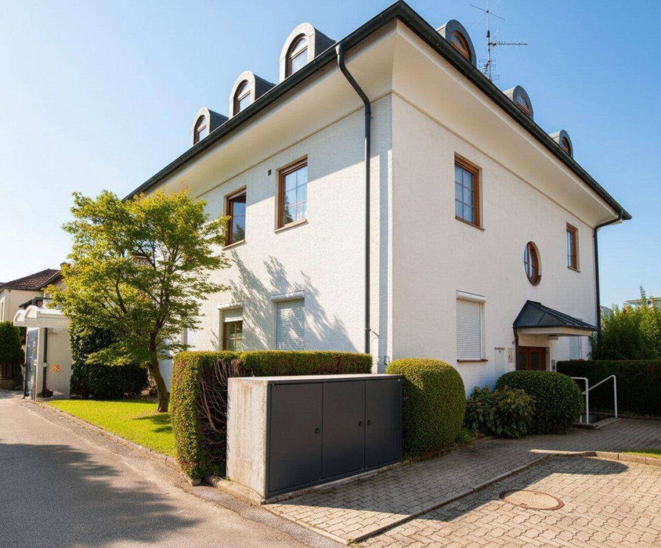Modernes Wohnhaus mit weißer Fassade, Mansarddach und gepflegter Hecke im Vorgarten.