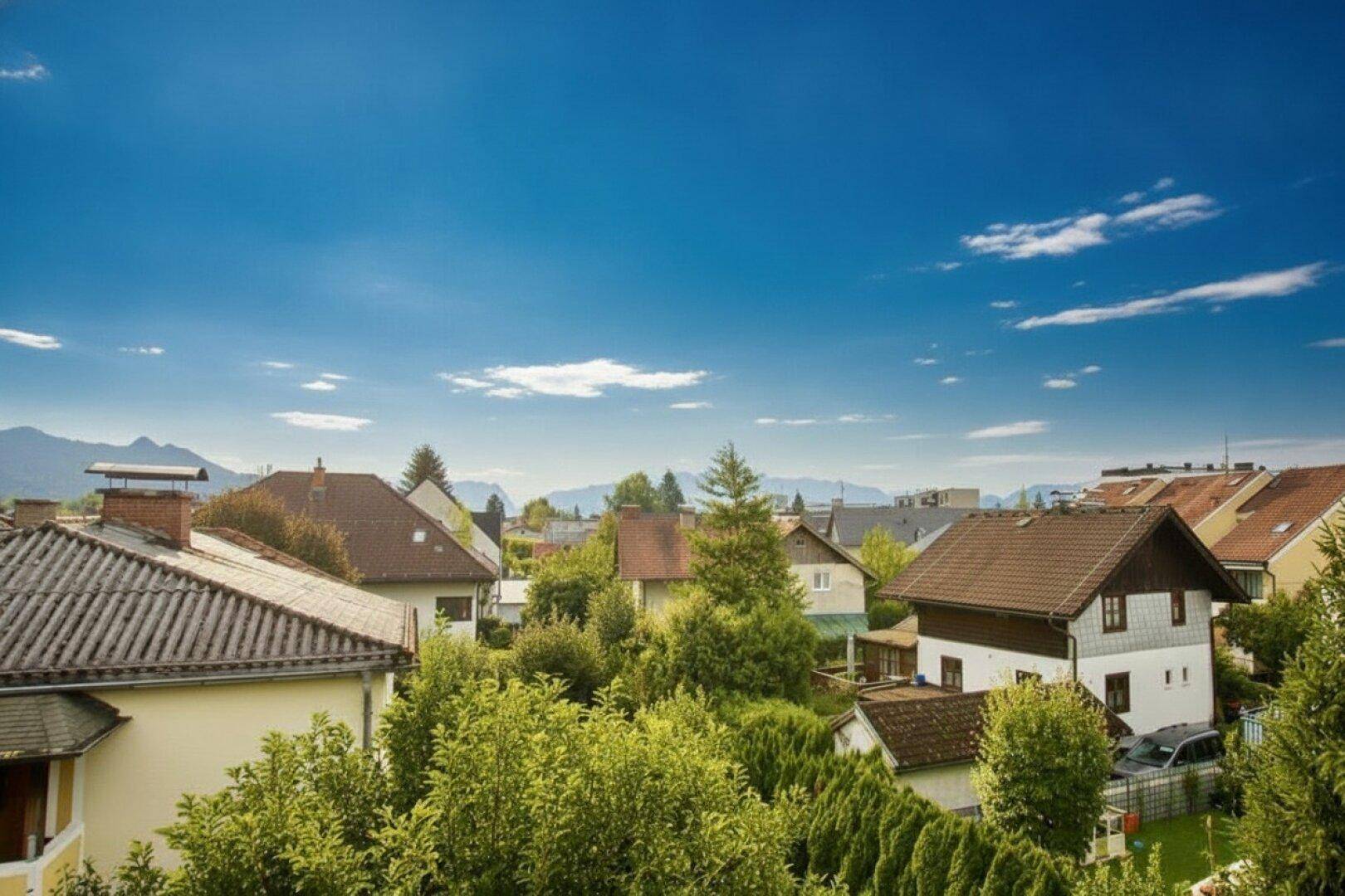 Weitläufiger Blick über eine Wohnsiedlung mit grünen Gärten und Bergen im Hintergrund.