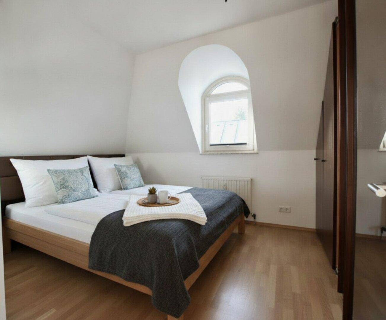 Gemütliches Schlafzimmer mit Holzbett, Parkettboden und charakteristischem Rundbogenfenster.
