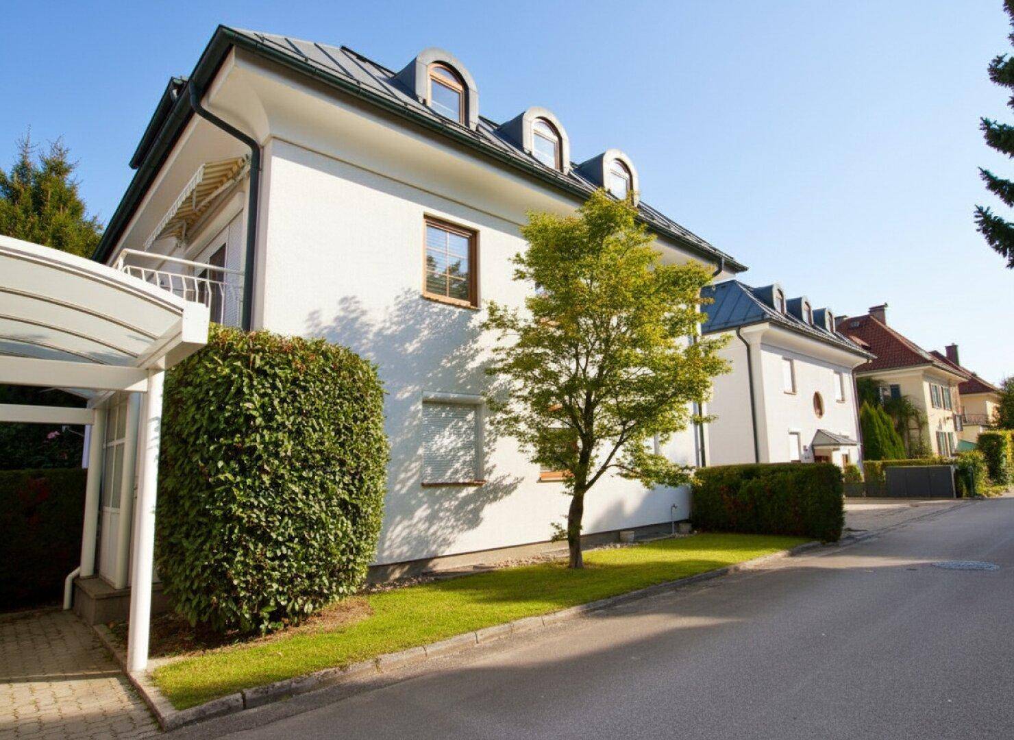Elegantes Wohnhaus mit weißer Fassade, Mansarddach und gepflegtem Vorgarten an einer ruhigen Straße.