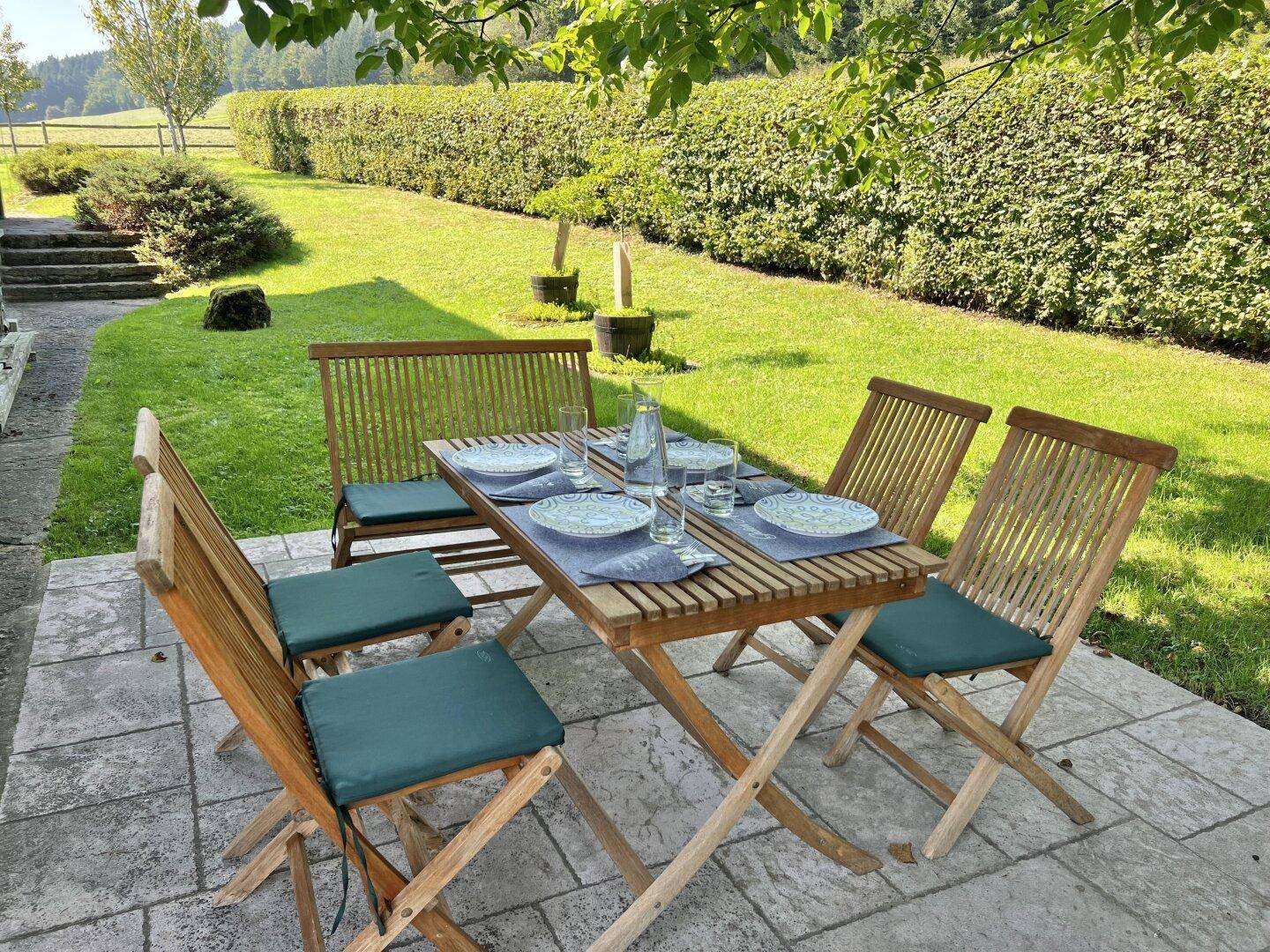 Sonnige Terrasse mit Holztisch und Stühlen, umgeben von gepflegtem Rasen und hohen Hecken.