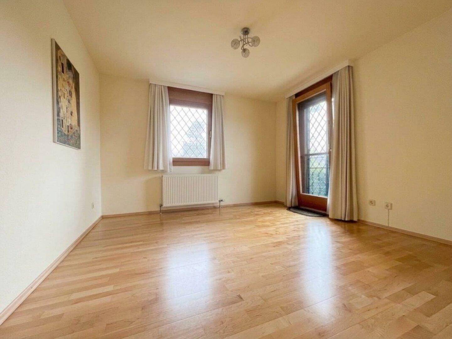 Leeres Zimmer mit hellem Parkettboden, Heizkörper und Zugang zu einem französischen Balkon.