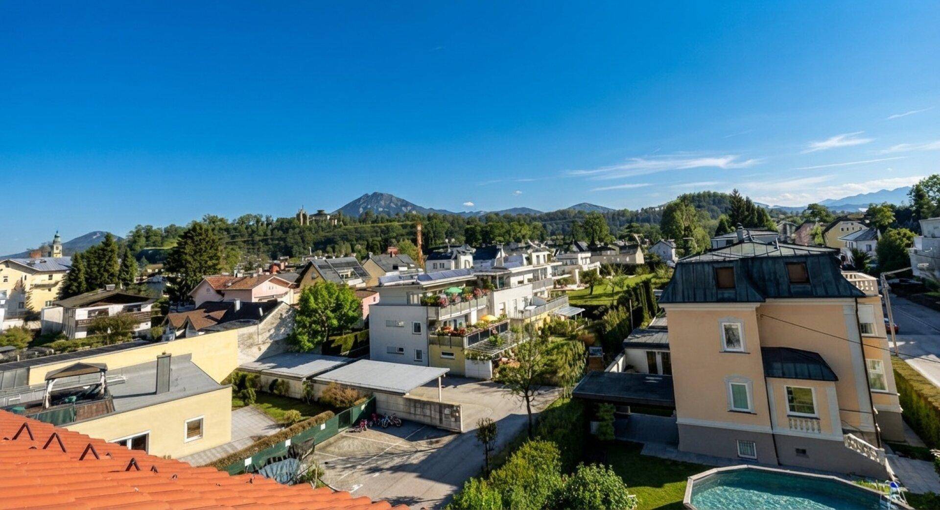 Panoramablick über eine Wohngegend mit Häusern, viel Grün und Bergen im Hintergrund.