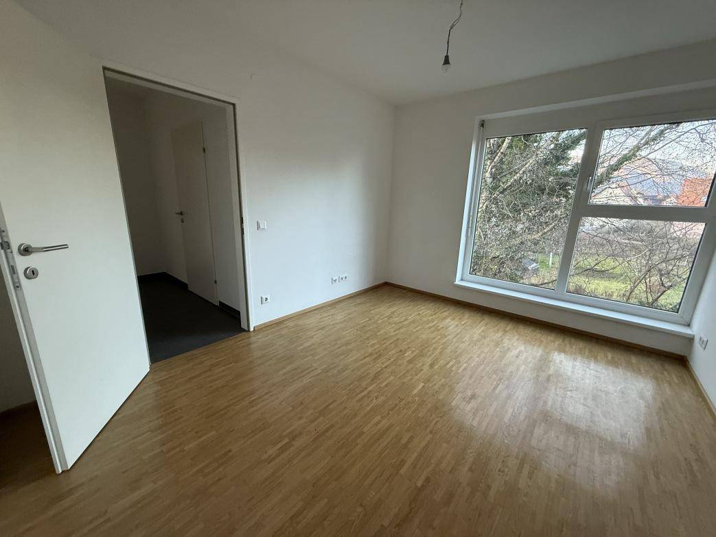 Helles Schlafzimmer mit Parkettboden und einem großen Fenster, das viel Tageslicht hereinlässt.