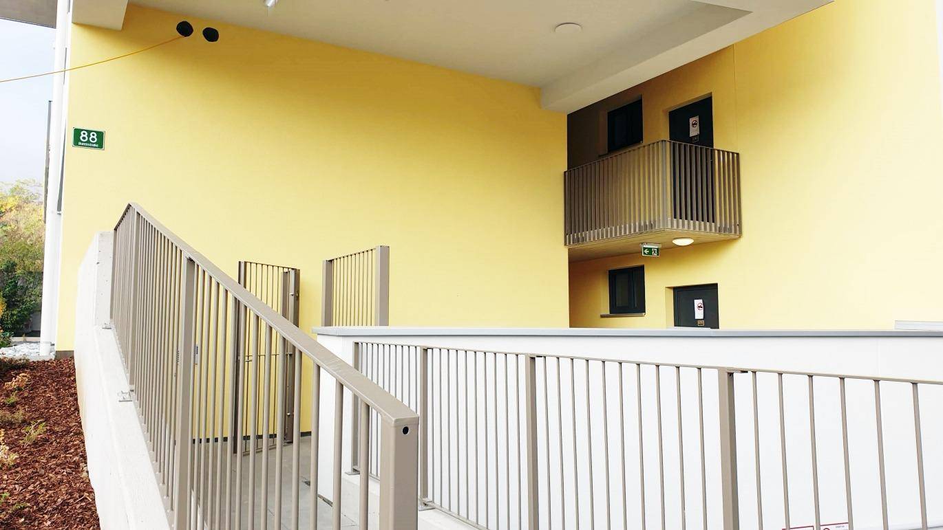 Moderner Eingangsbereich des Gebäudes mit gelber Fassade und einer barrierefreien Rampe.