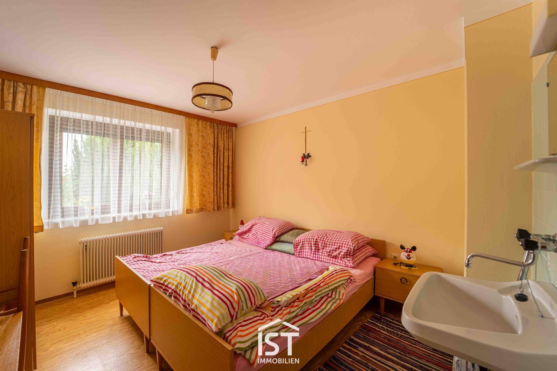 Helles Schlafzimmer mit Doppelbett, Waschbecken und Fenster mit Vorhängen.