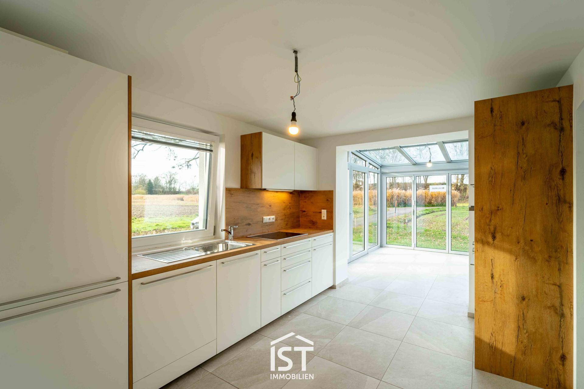 Moderne, leere Küche mit weißen Schränken und Holzakzenten, mit Blick in den Wintergarten.