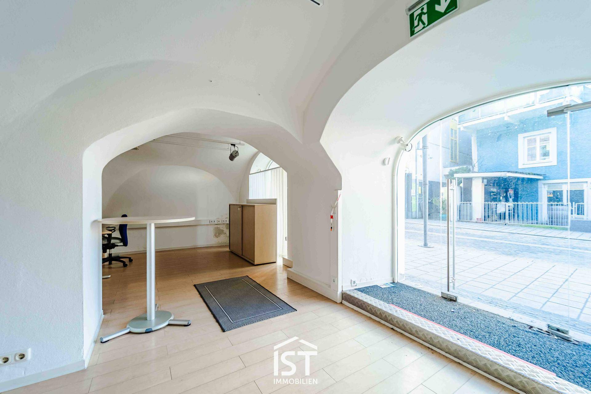 Heller Büroraum mit hohen Gewölbedecken, Holzboden und großen Schaufenstern zur Straße hin, die viel Licht hereinlassen.