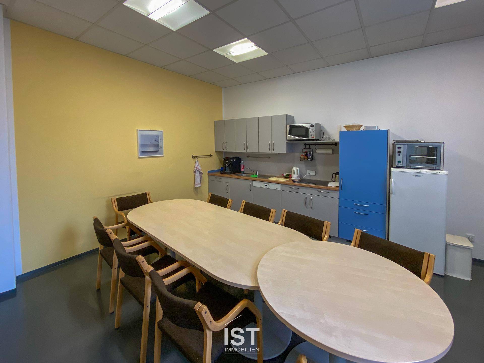 Funktionale Teeküche mit langer Tafel, grauen Schränken und verschiedenen Küchengeräten für Mitarbeiter.