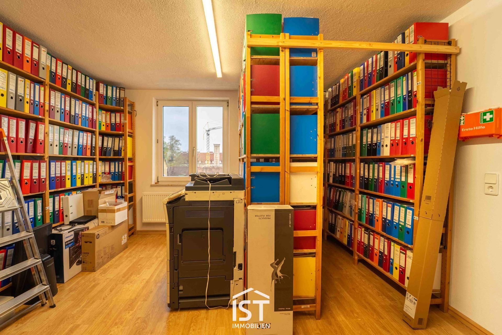 Archivraum mit Regalen voller Aktenordner, einem Fenster und einem Drucker, ideal für die Dokumentenverwaltung.