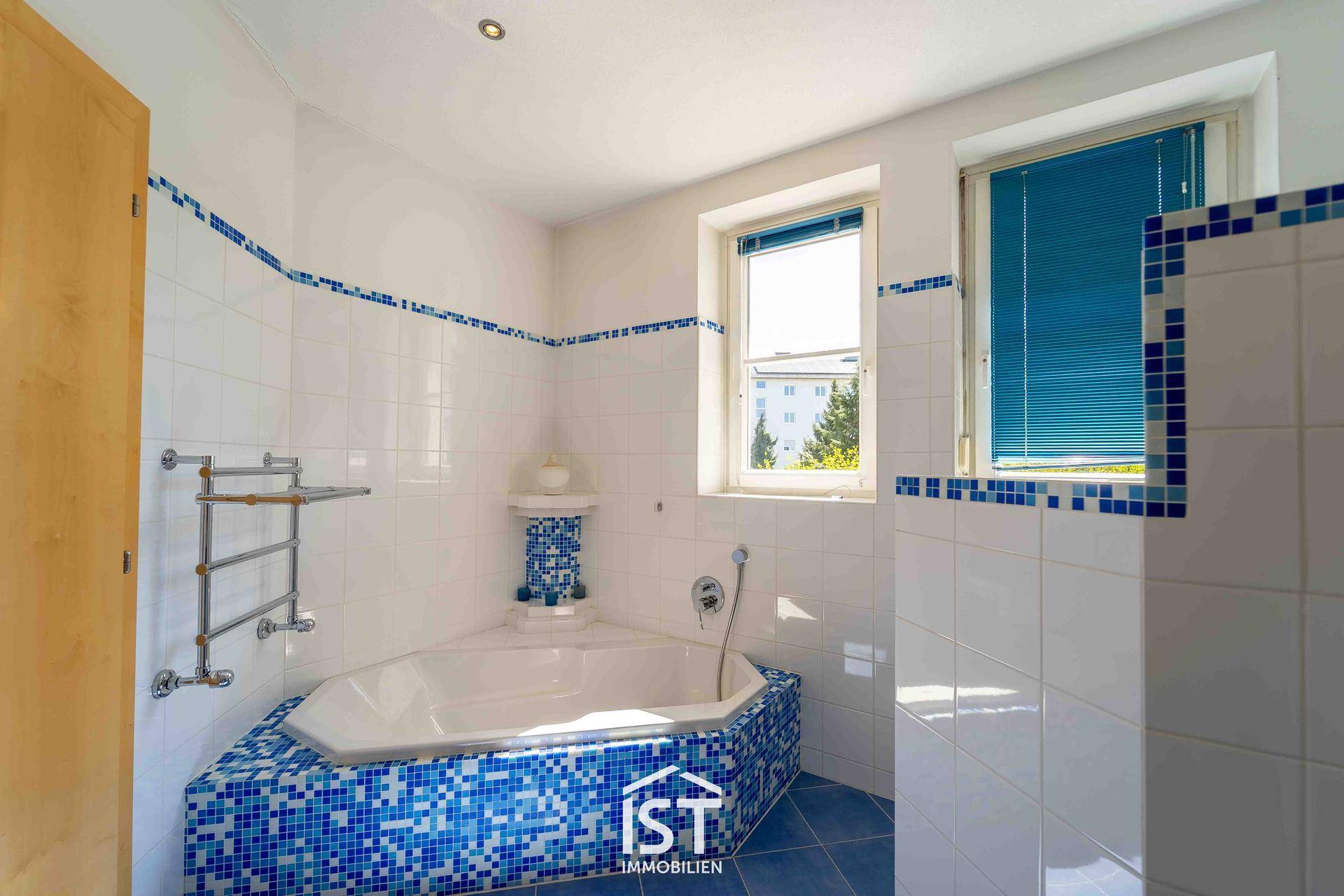Badezimmer mit Eckbadewanne, blauen Mosaikfliesen und Fenster für natürliche Belüftung.