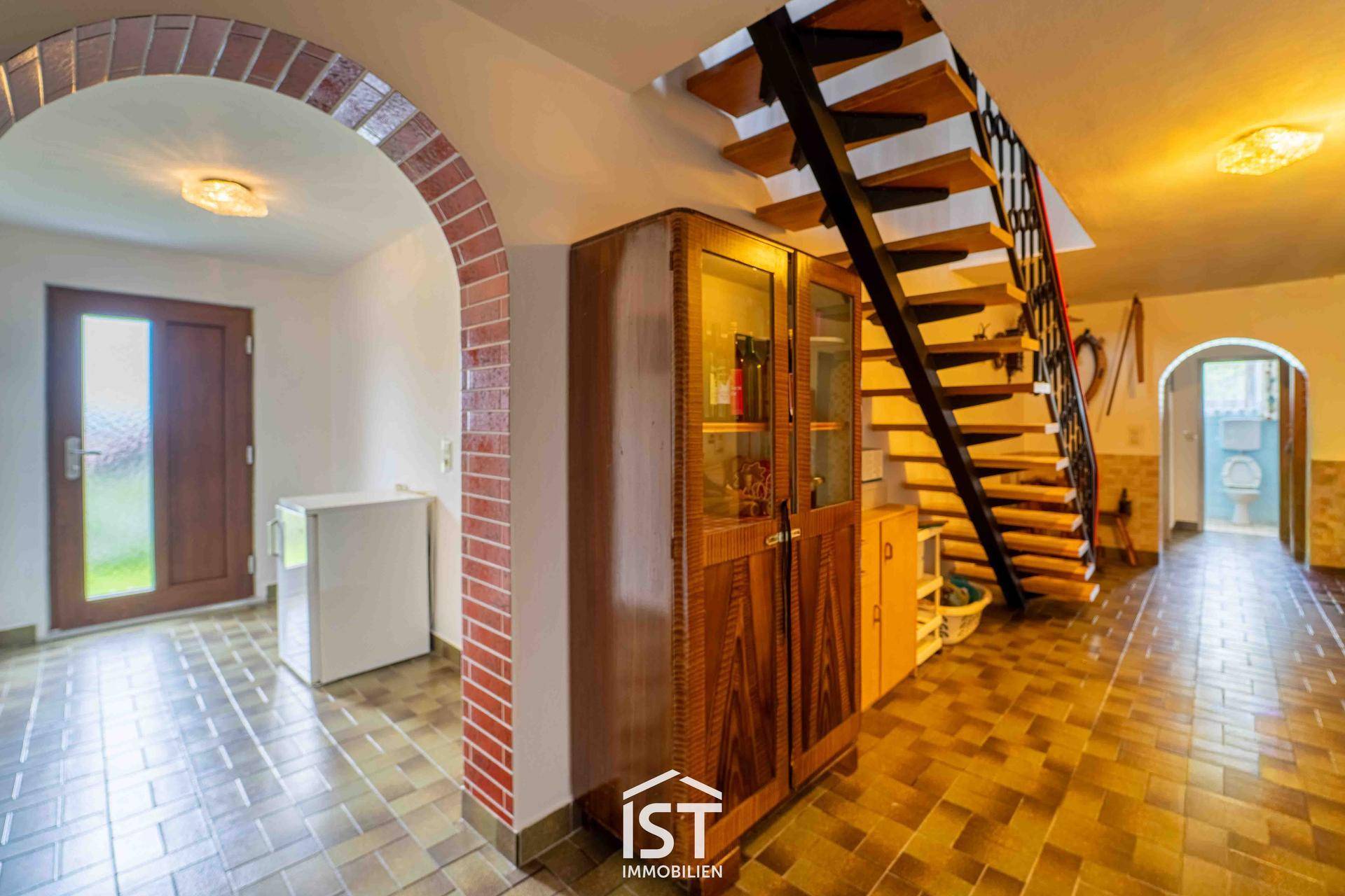 Eingangsbereich mit Ziegelbogen, gefliestem Boden und offener Holztreppe.