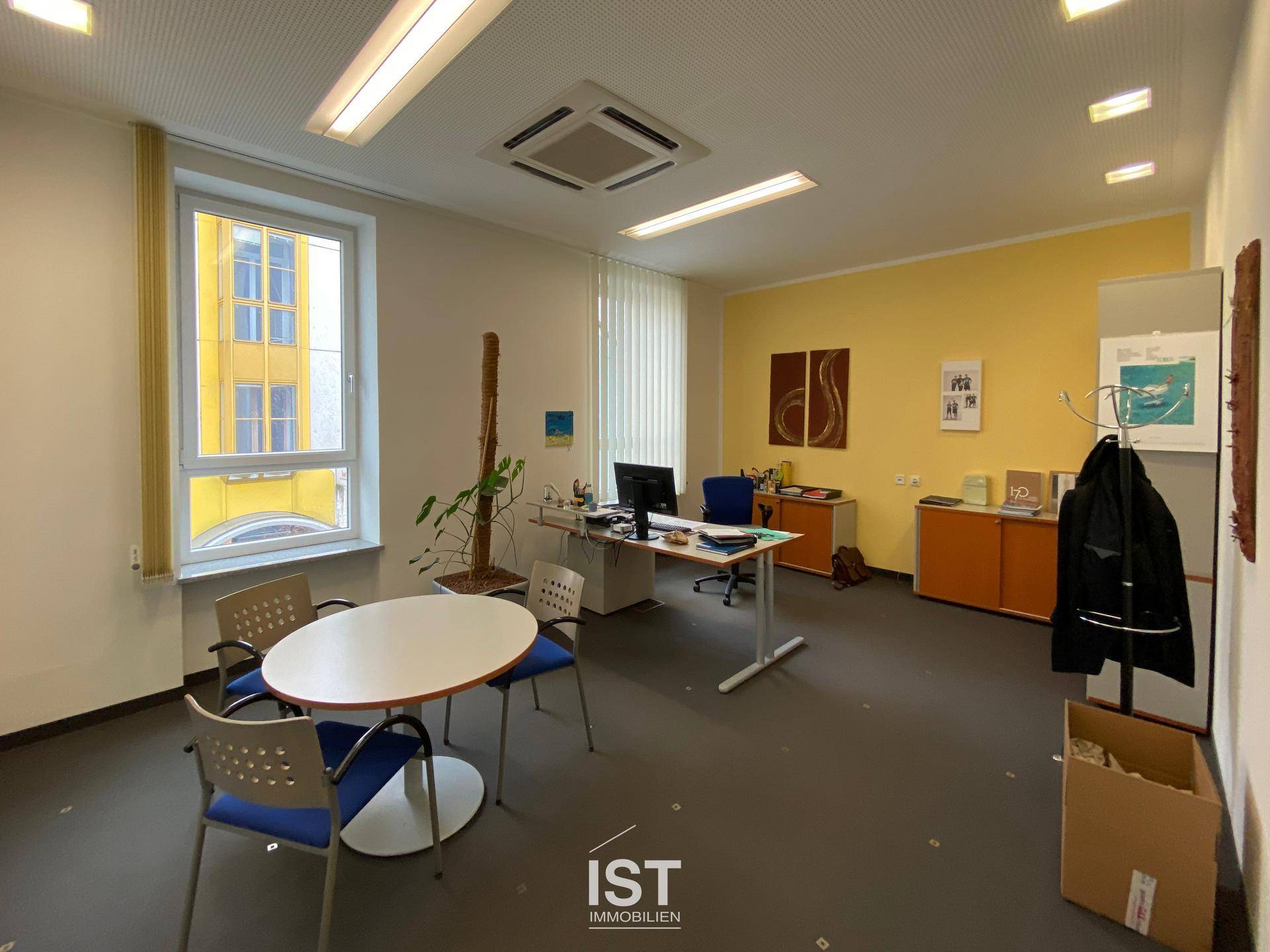 Helles Einzelbüro mit großem Fenster, gelber Akzentwand und mehreren Arbeitsplätzen in einem modernen Stil.