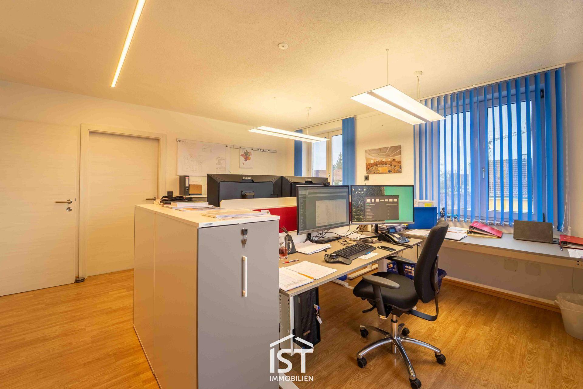 Geräumiges Büro mit mehreren Arbeitsplätzen, ausgestattet mit Computern und blauen Jalousien an den Fenstern.