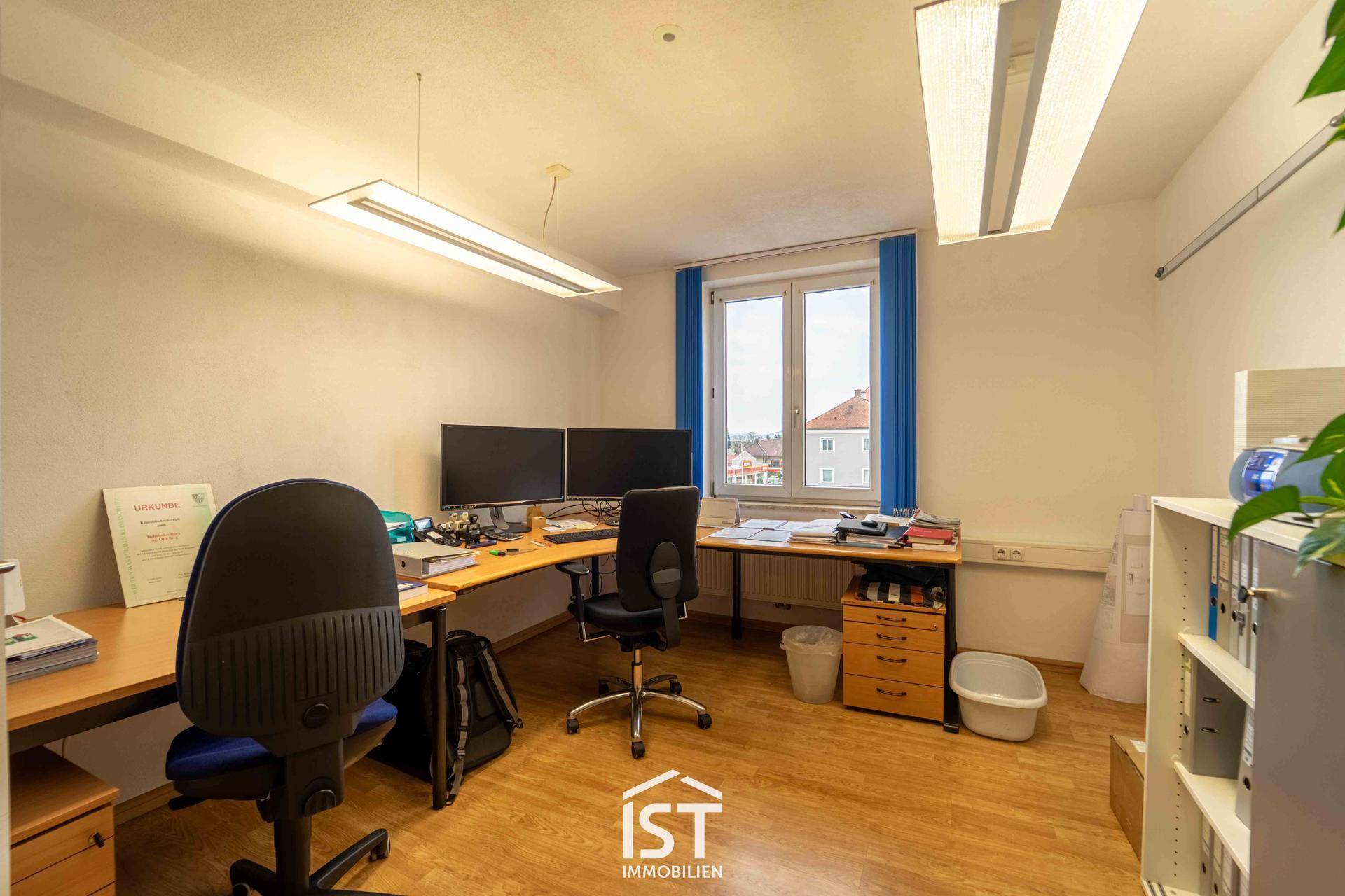 Helles Büro mit zwei Arbeitsplätzen, großen Fenstern und Blick auf die Umgebung, ausgestattet mit Büromöbeln.