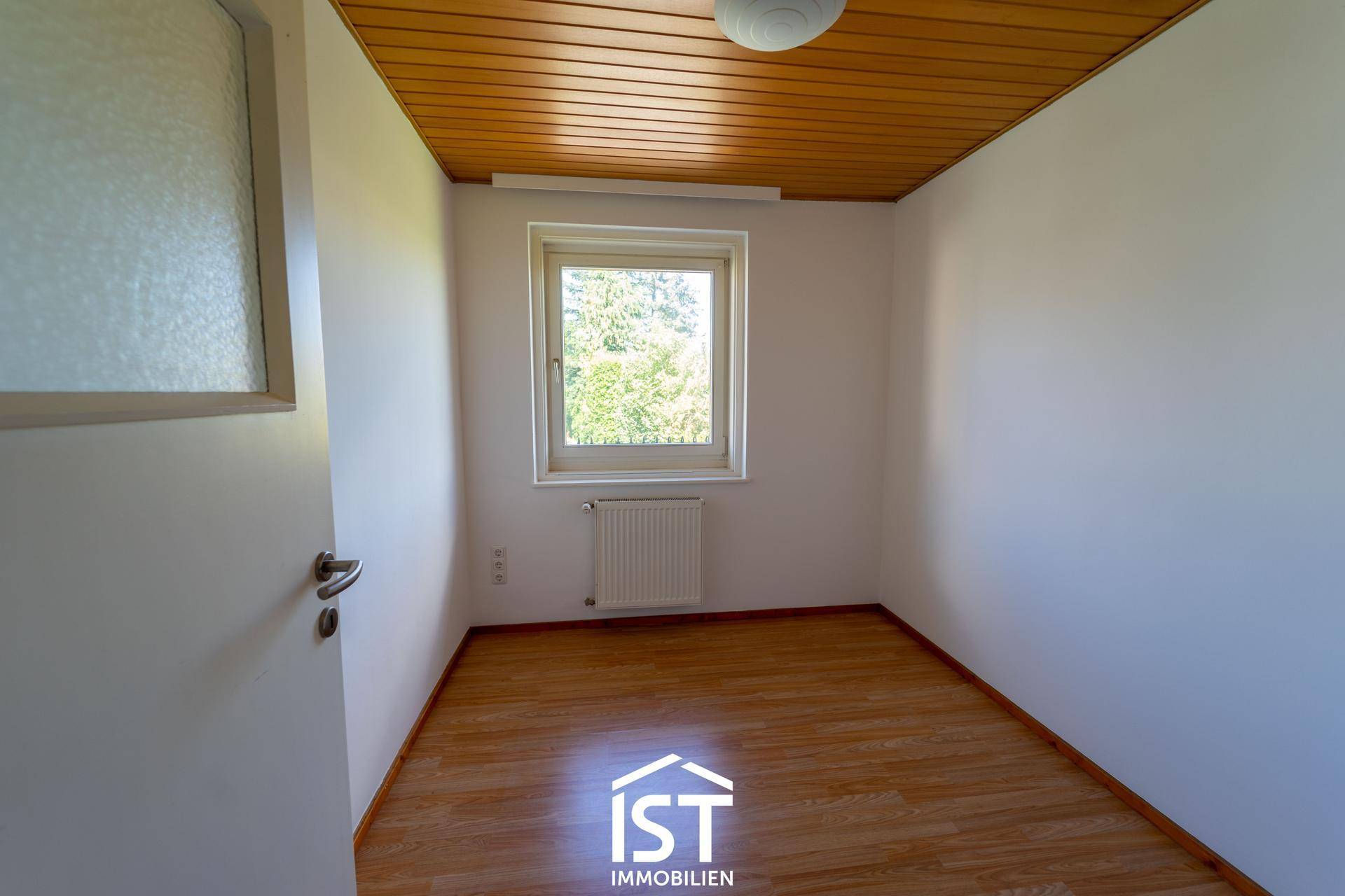 Kleines Zimmer mit Laminatboden, Holzdecke und Fenster mit Blick ins Grüne.