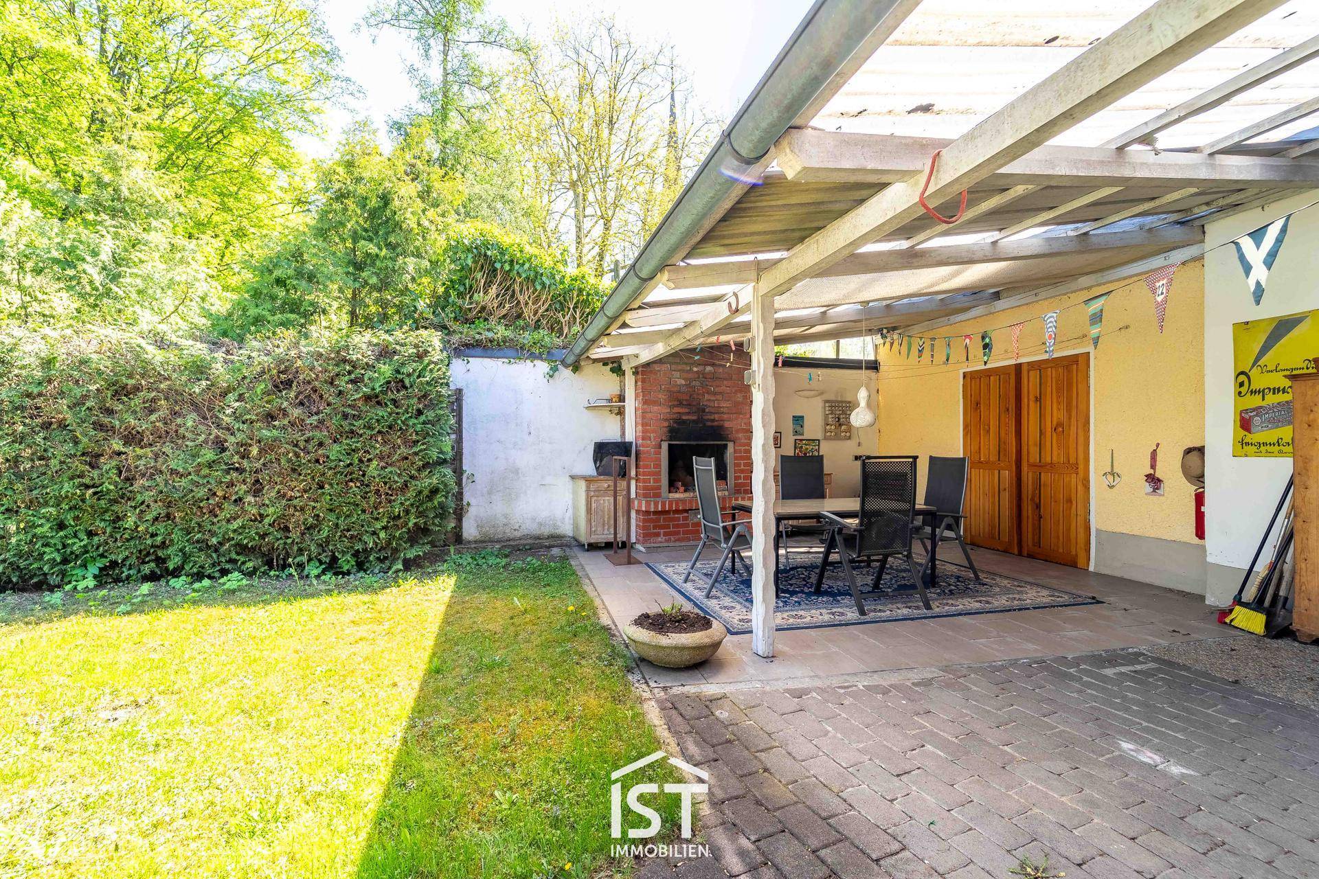 Überdachte Terrasse mit Grillplatz und Gartenmöbeln, umgeben von grüner Hecke.