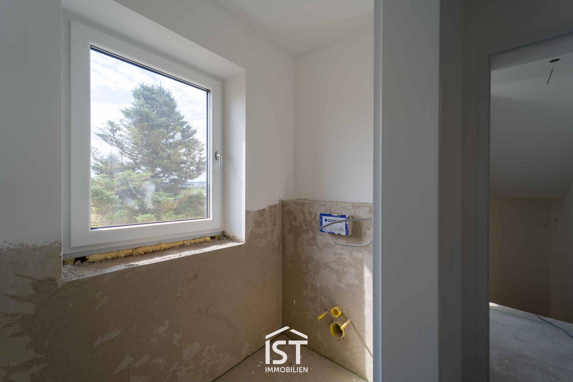 Rohbau eines Badezimmers mit vorbereiteten Anschlüssen für Toilette und Waschbecken sowie einem Fenster.