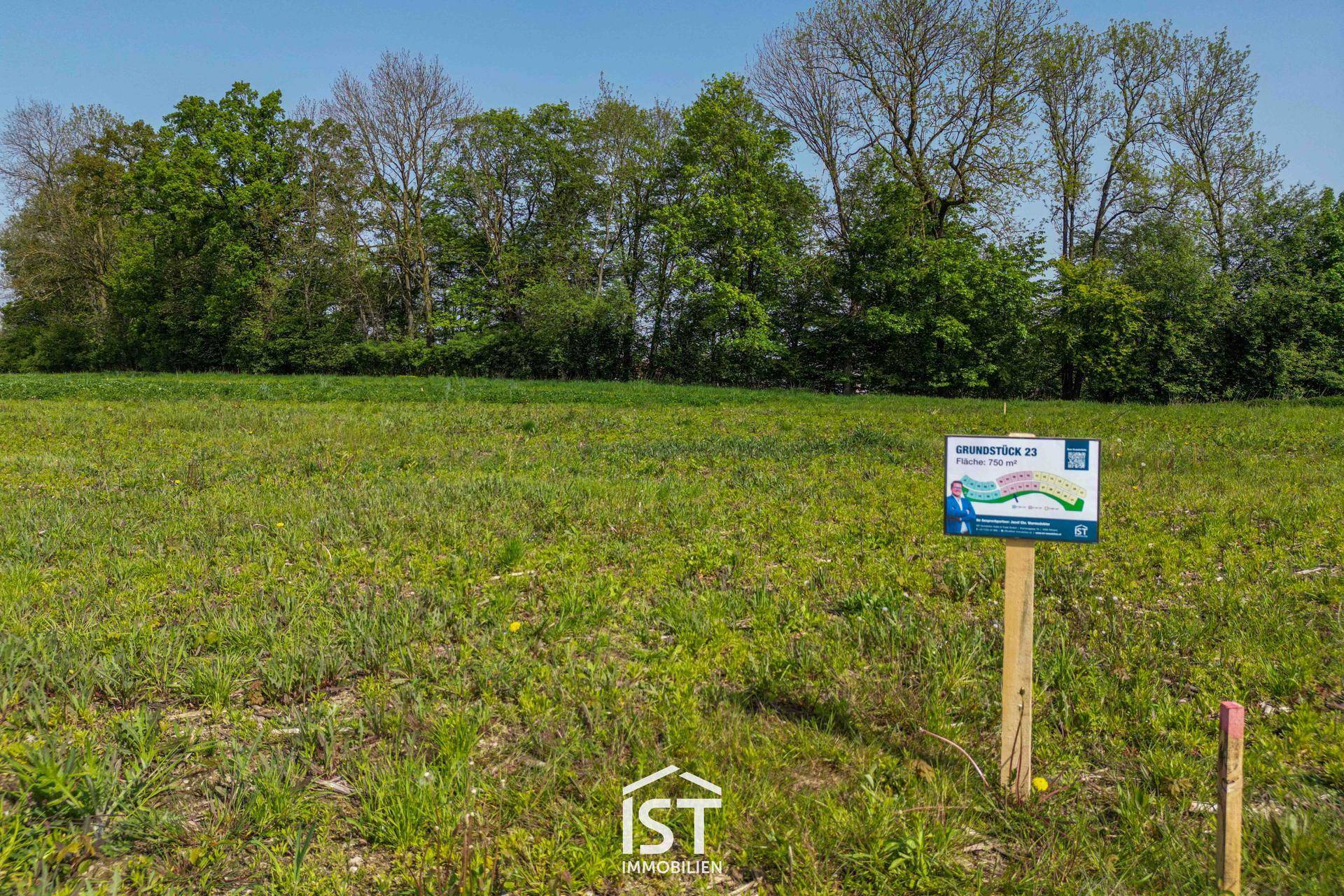 Schild für Grundstück 23 auf einer grünen Wiese mit Bäumen im Hintergrund unter blauem Himmel.