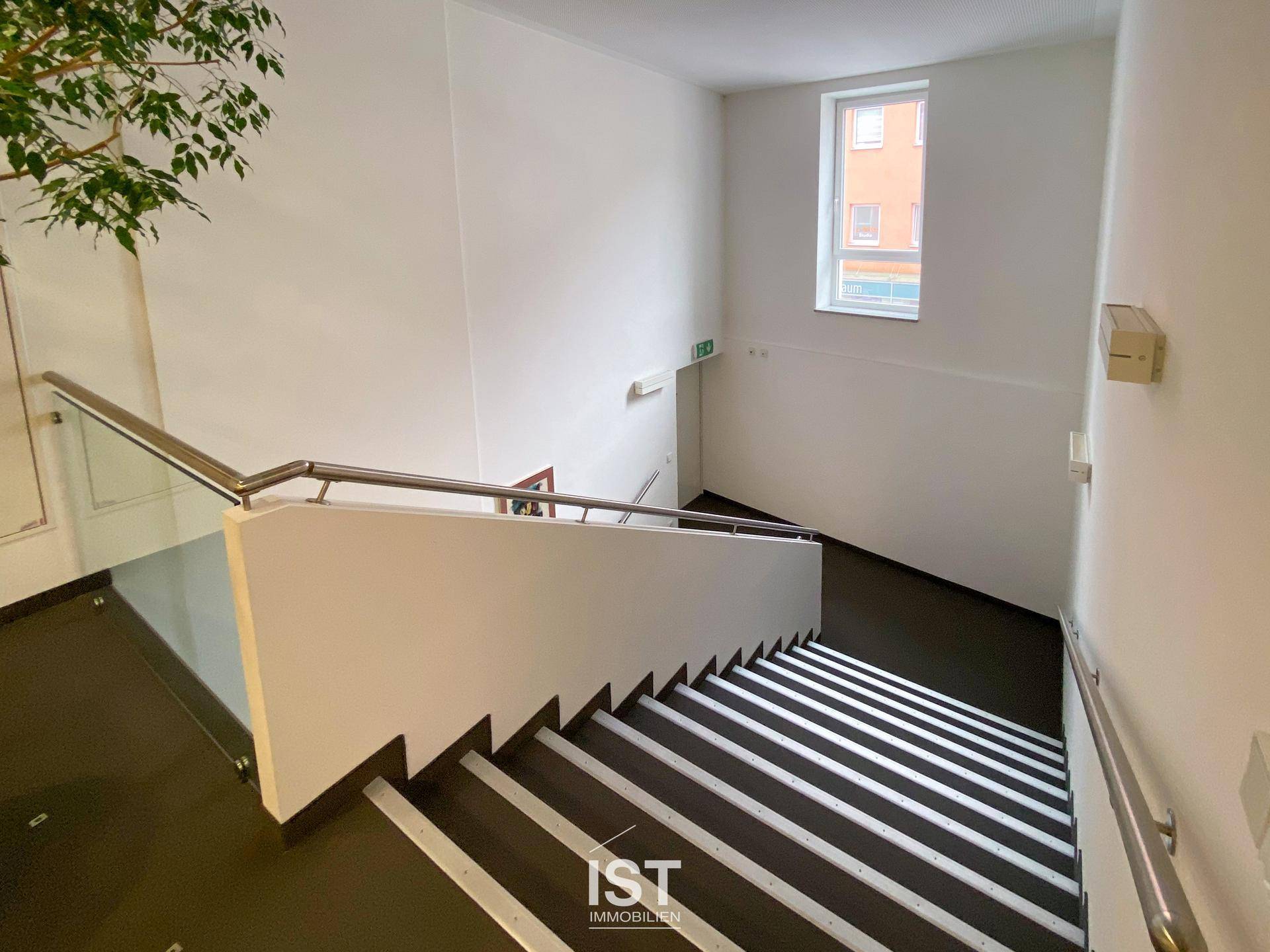 Moderner Treppenaufgang mit Edelstahlgeländer, breiten Stufen und einem Fenster, das Tageslicht hereinlässt.