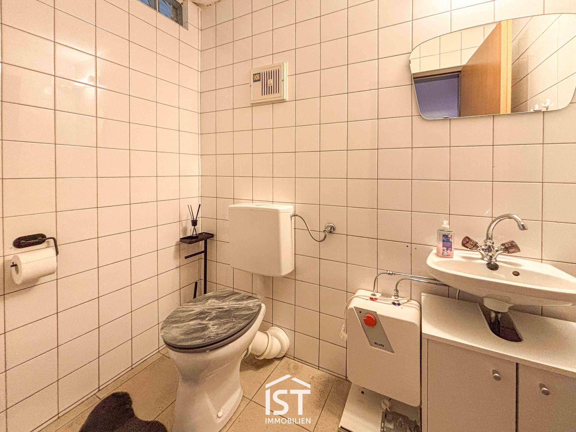 Einfache Toilette mit weißen Fliesen an Wänden und Boden, ausgestattet mit Waschbecken und WC.