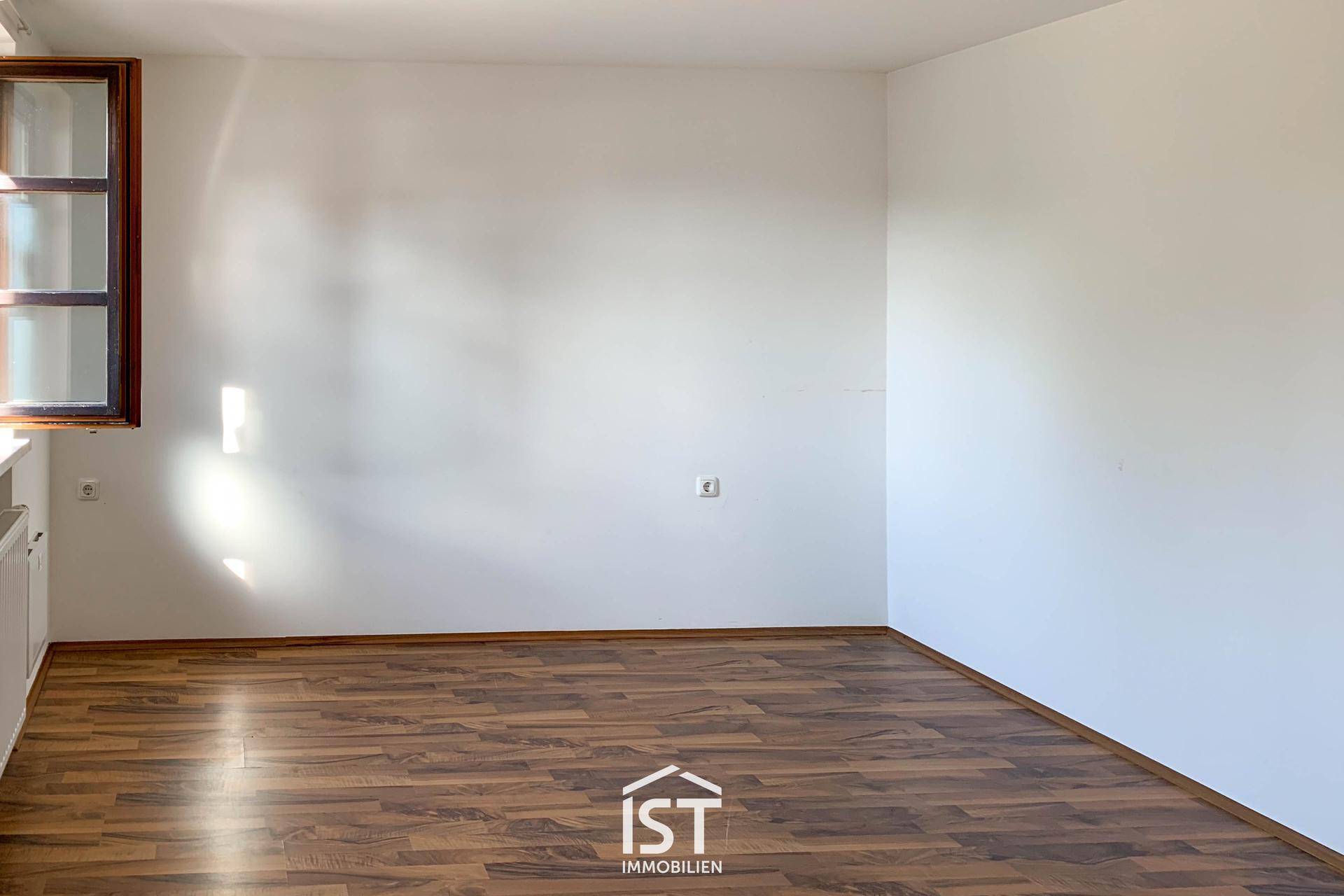 Leeres Zimmer mit hellem Laminatboden und einem Fenster, das Tageslicht hereinlässt.