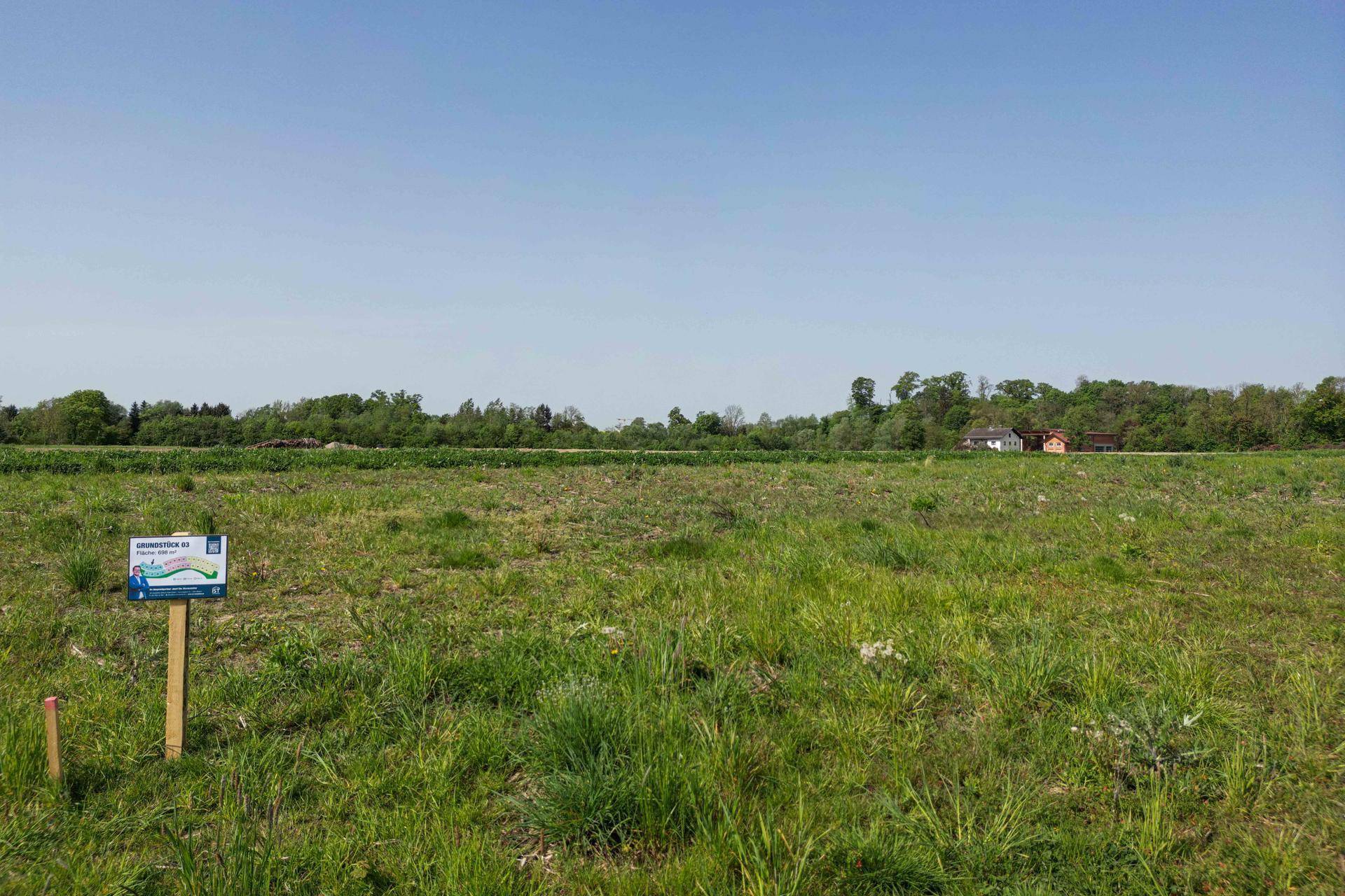 Weitläufige grüne Wiese mit einem kleinen Hinweisschild und Bäumen am Horizont unter blauem Himmel.