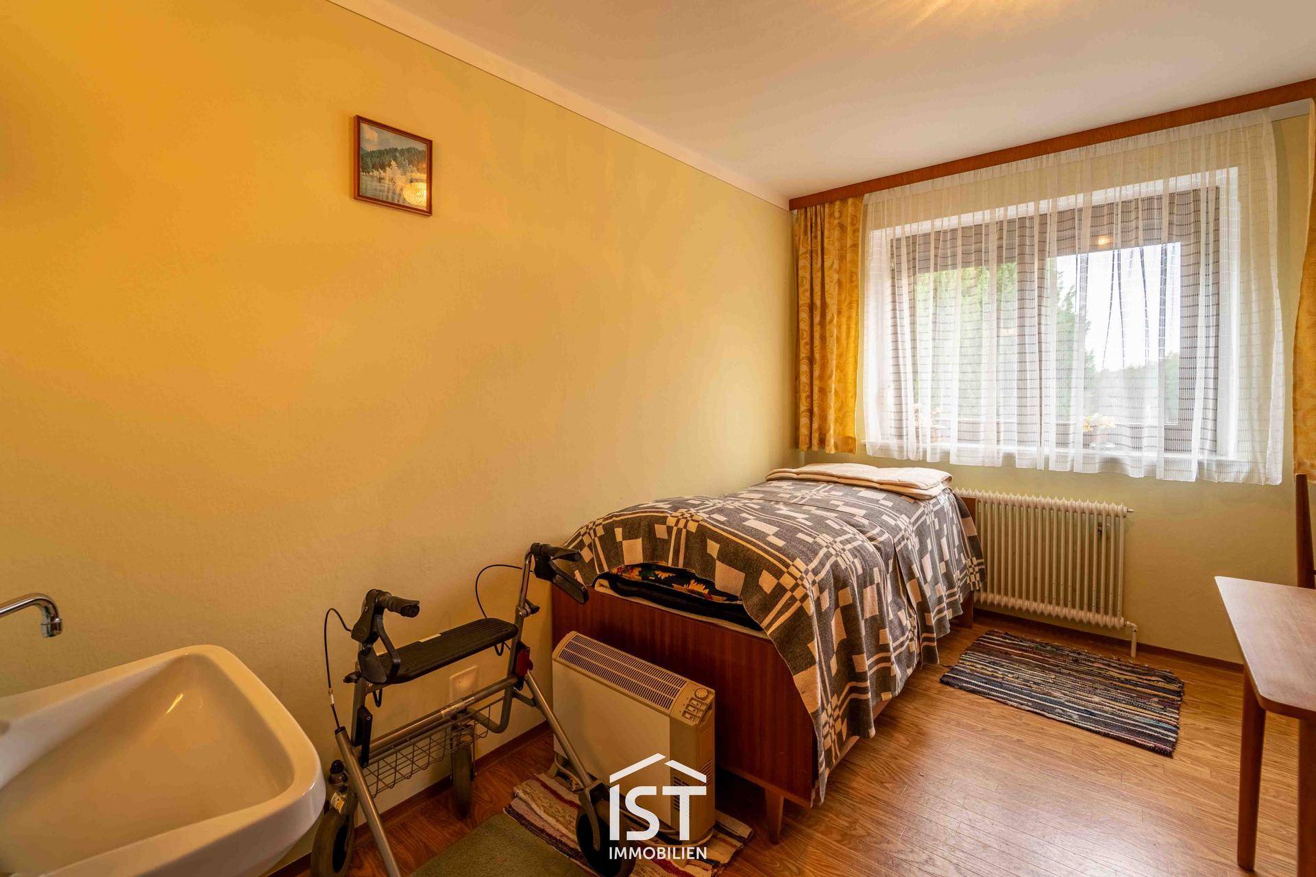 Schlafzimmer mit Einzelbett, Waschbecken und Heizkörper unter dem Fenster.