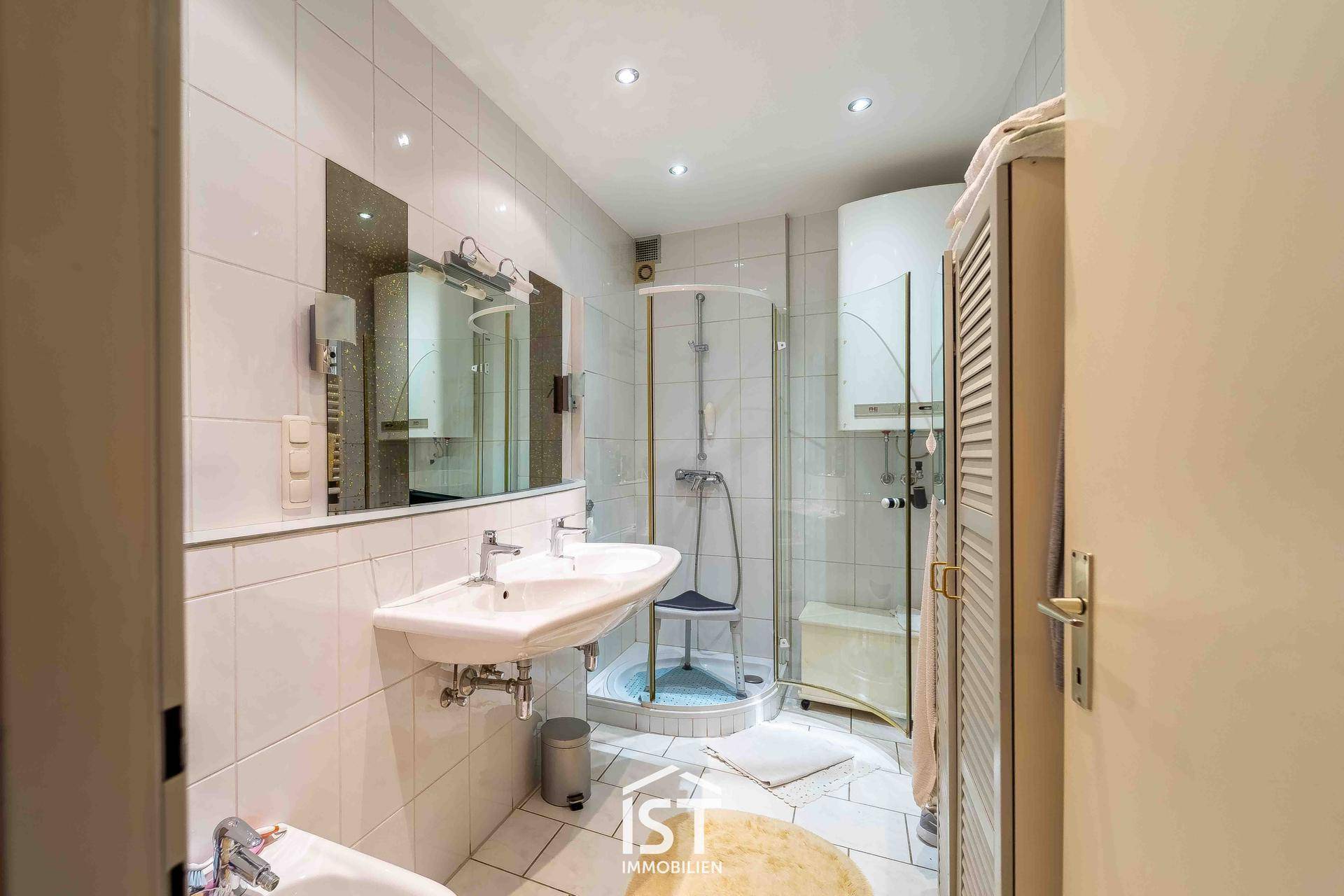 Gefliestes Badezimmer mit Doppelwaschbecken, Dusche und weißer Einrichtung.