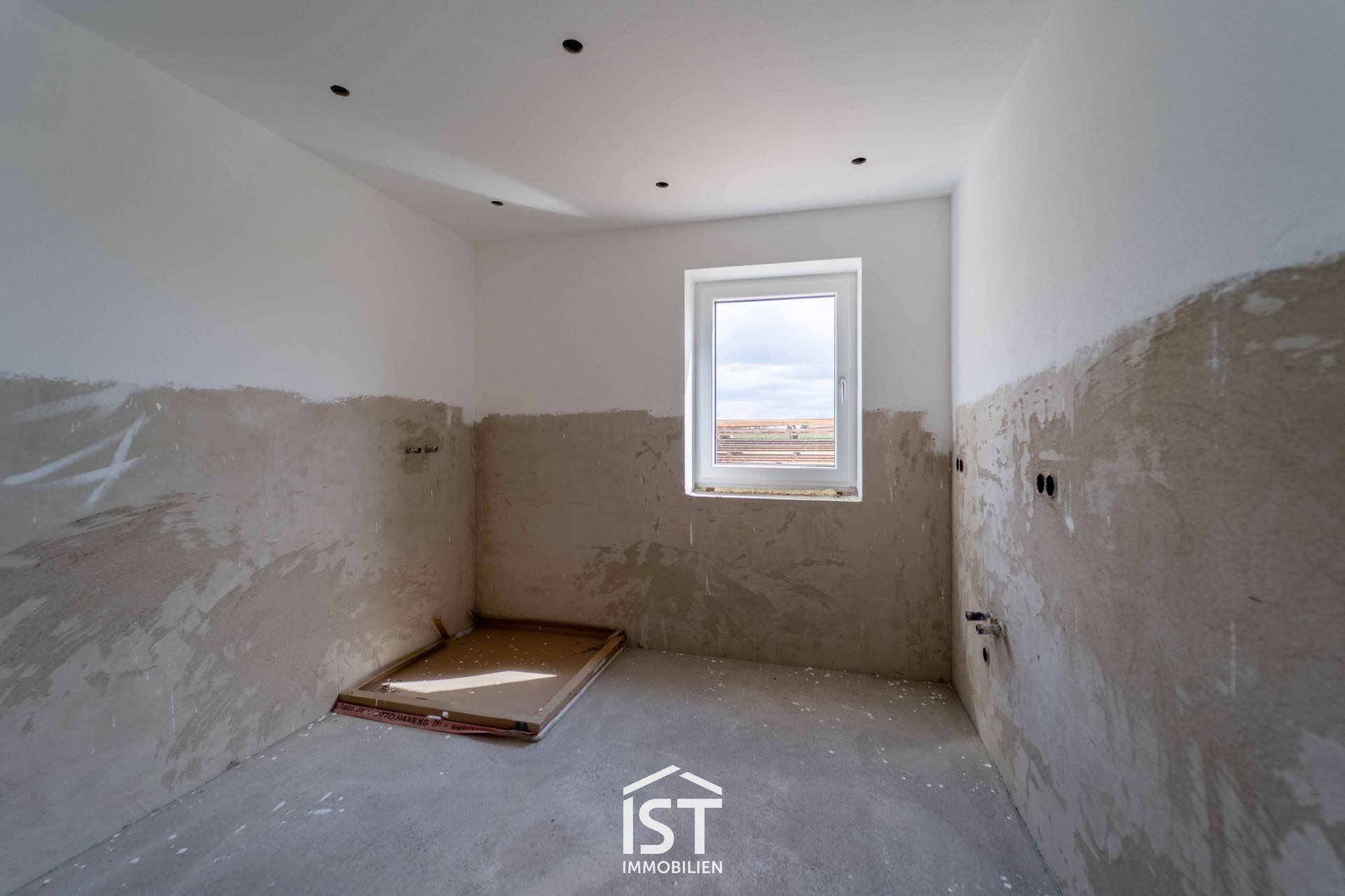 Unfertiges Badezimmer mit Fenster und vorbereiteten Anschlüssen für Sanitärinstallationen.