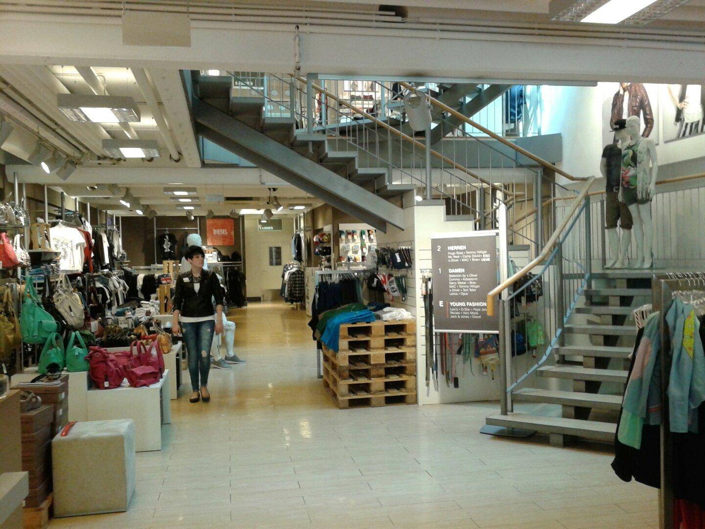 Großer Verkaufsraum mit Kleidung, Accessoires und einer zentralen Treppe, die zu weiteren Etagen führt. Die Einrichtung wirkt modern und funktional.