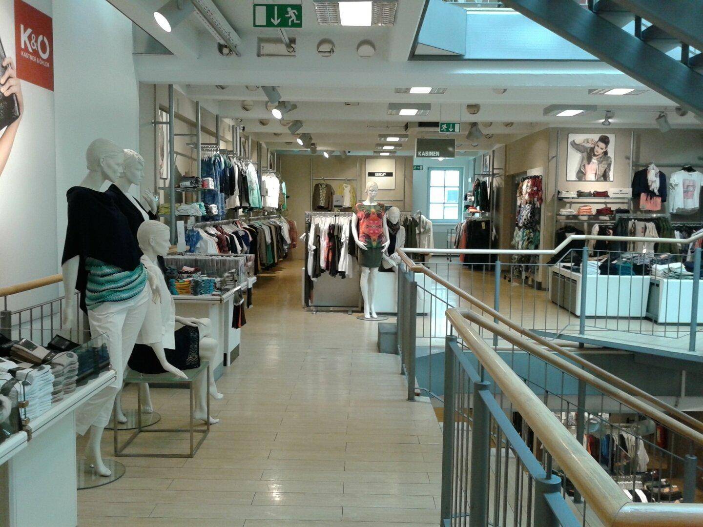 Weitläufiger Verkaufsbereich mit Schaufensterpuppen, Kleiderständern und Zugang zu weiteren Etagen über eine Treppe.